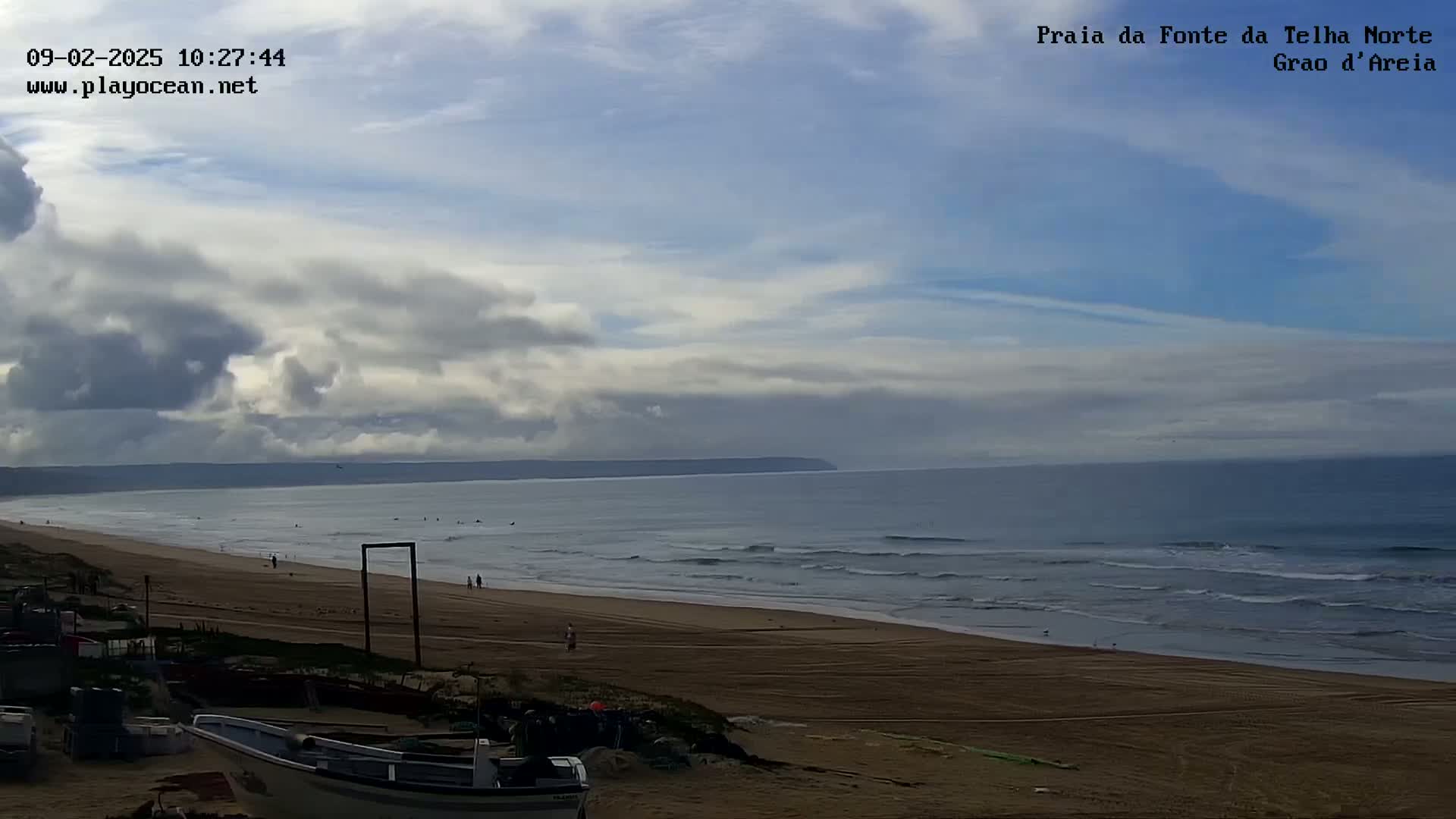 A mostly cloudy sky overlooks a long sandy beach with a calm ocean and a few people walking along the shore.