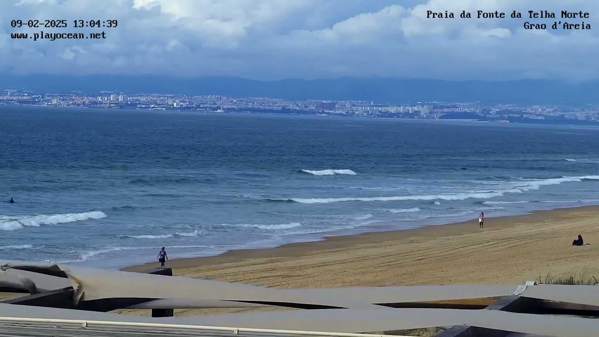 A sandy beach with gently rolling waves under a partly cloudy sky overlooks a distant cityscape.