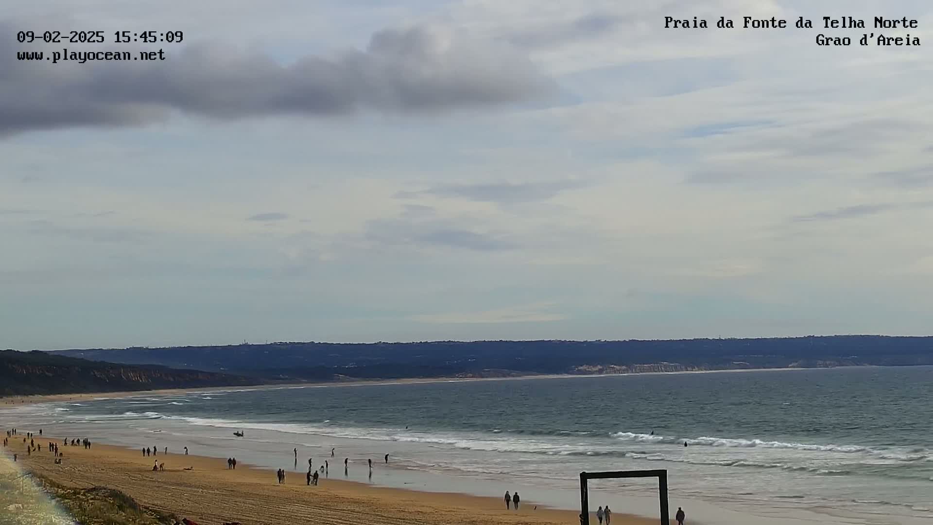 A sandy beach with scattered people stretches along a calm ocean under a mostly cloudy sky.