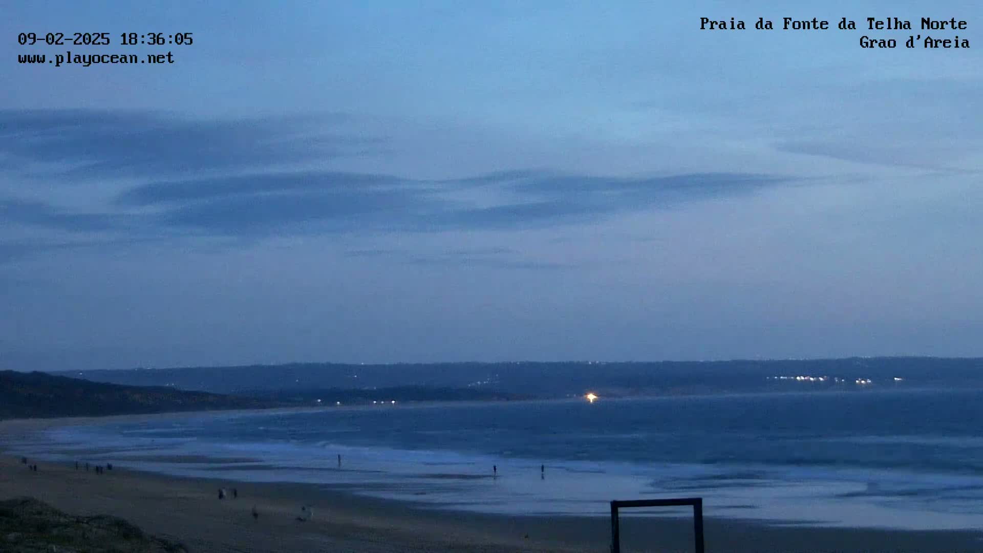 A twilight beach scene shows a sandy shore with a few people, gentle waves lapping the sand, and a dark, calm ocean under a mostly clear, dusky blue sky.