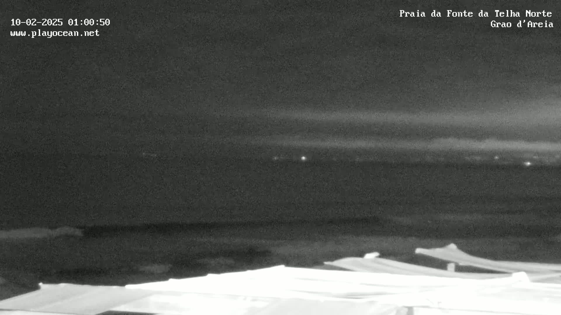 A nighttime view of a dark ocean under a cloudy sky, with several faint lights visible in the distance and covered structures in the foreground.