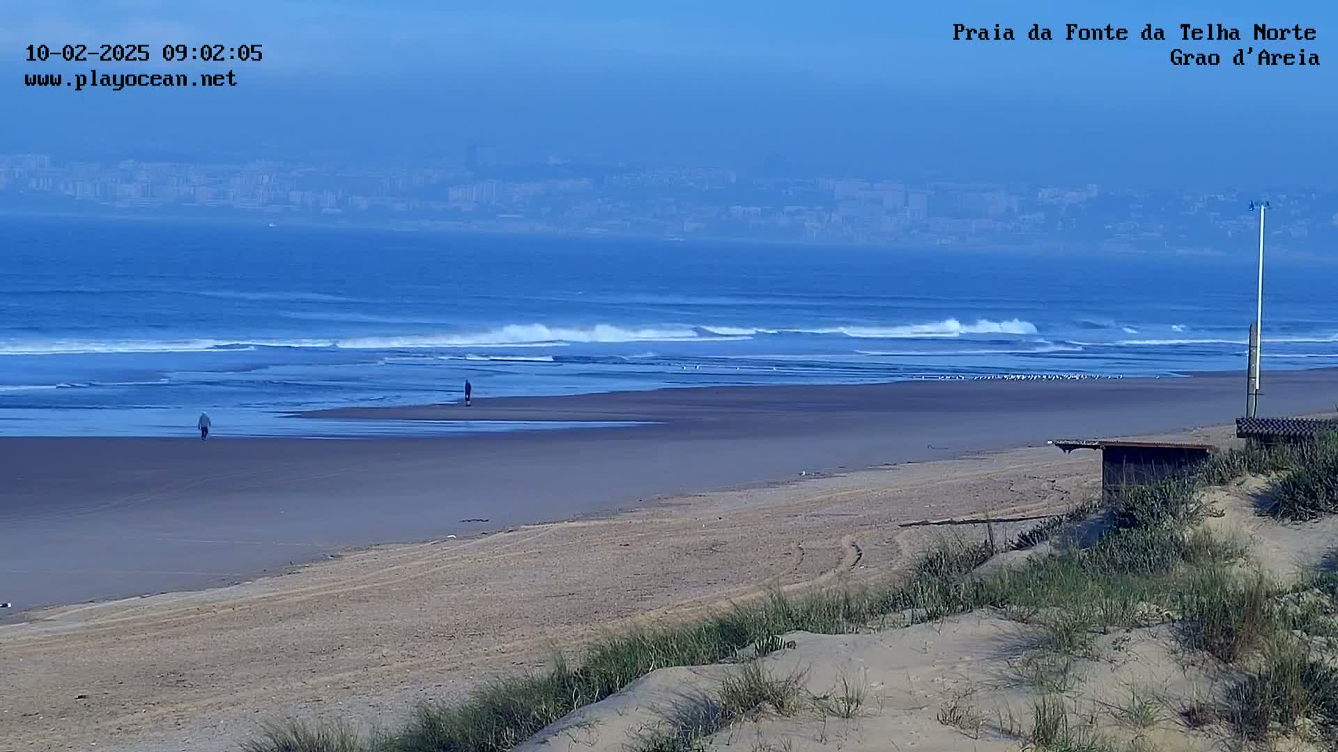 A sandy beach with a few people walking on it, gentle waves rolling into shore under a clear, hazy sky, and a distant city visible on the horizon.