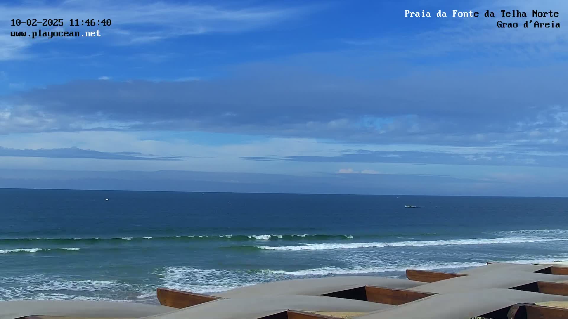 A mostly sunny day at a beach shows a calm ocean with small waves breaking on the shore, where several wooden structures are situated on the sand.