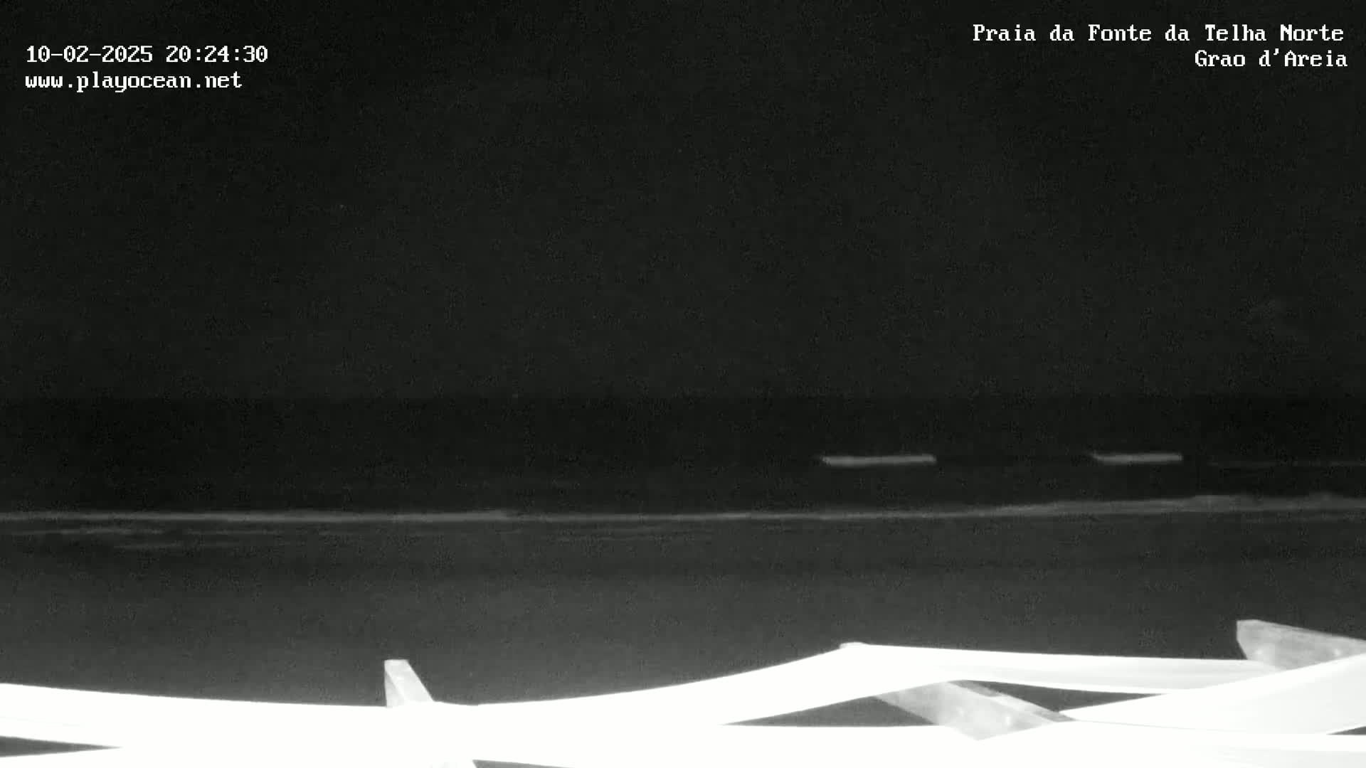 A nighttime, low-light view of a dark ocean shoreline with a white railing in the foreground.