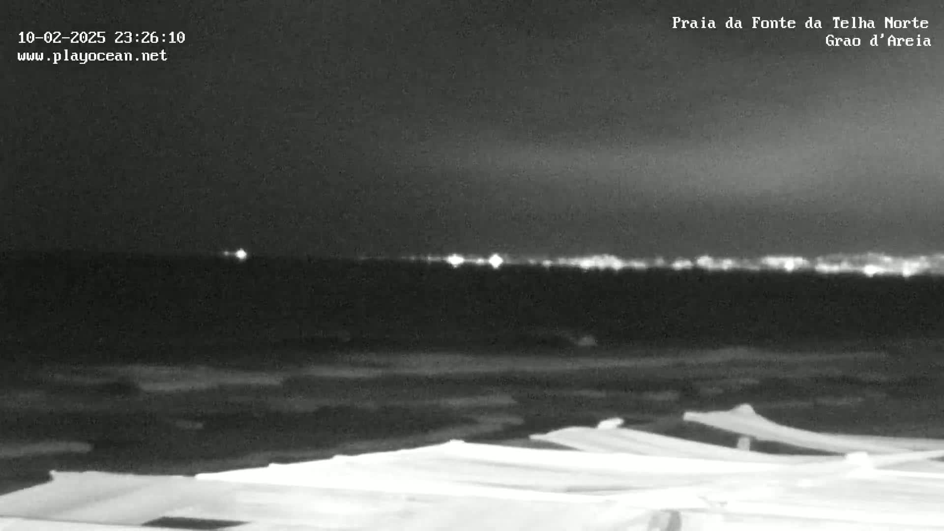 A nighttime, low-light image shows a dark beach with rows of covered objects in the foreground and a distant city skyline faintly illuminated on the horizon.