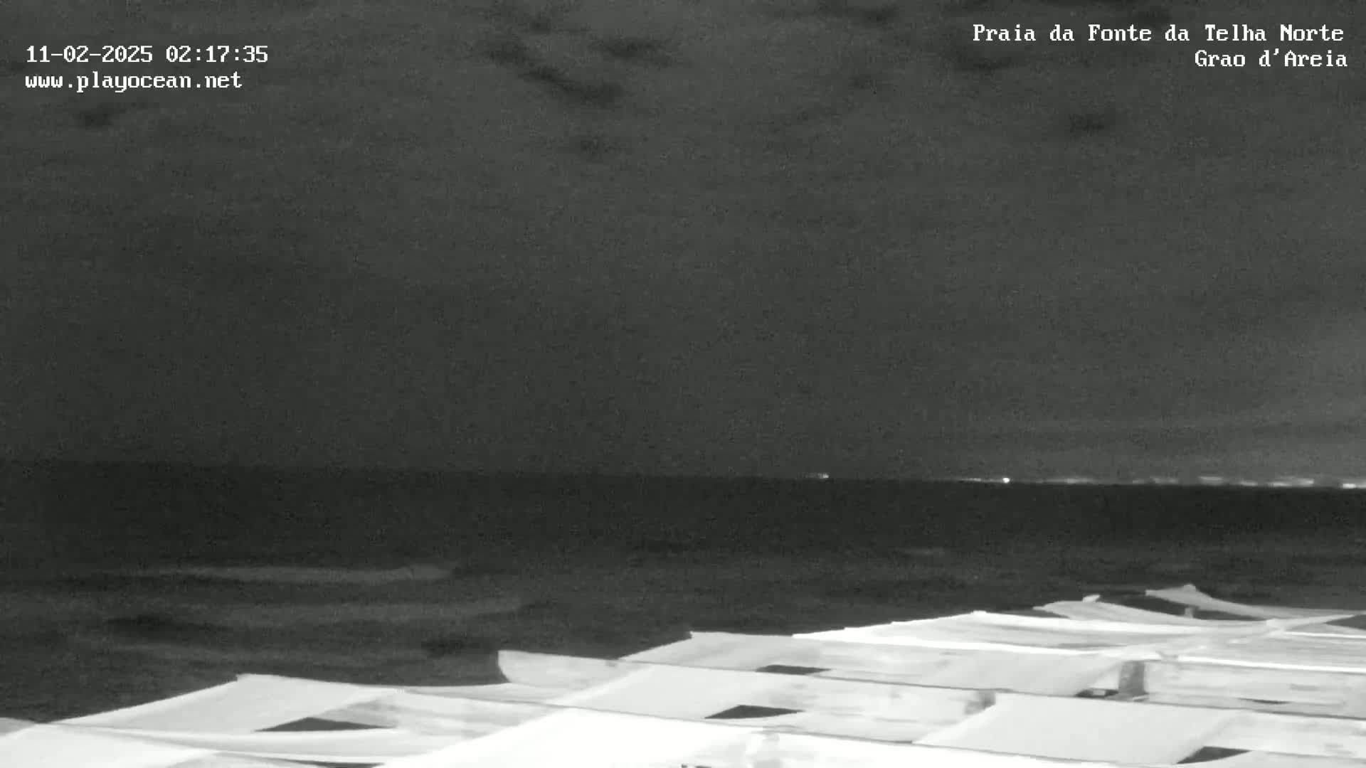 A nighttime beach scene shows a dark ocean under a cloudy sky, with rows of beach umbrellas in the foreground.