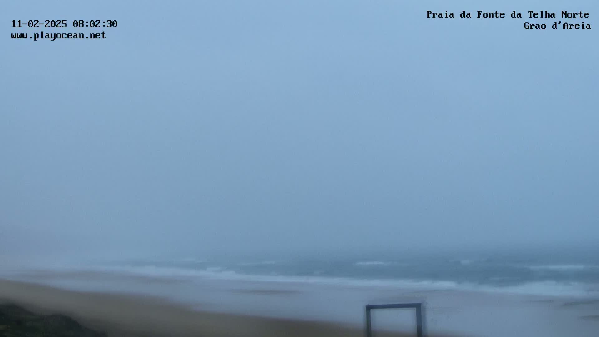 A foggy beach scene shows a sandy shore and a gray ocean with small waves.