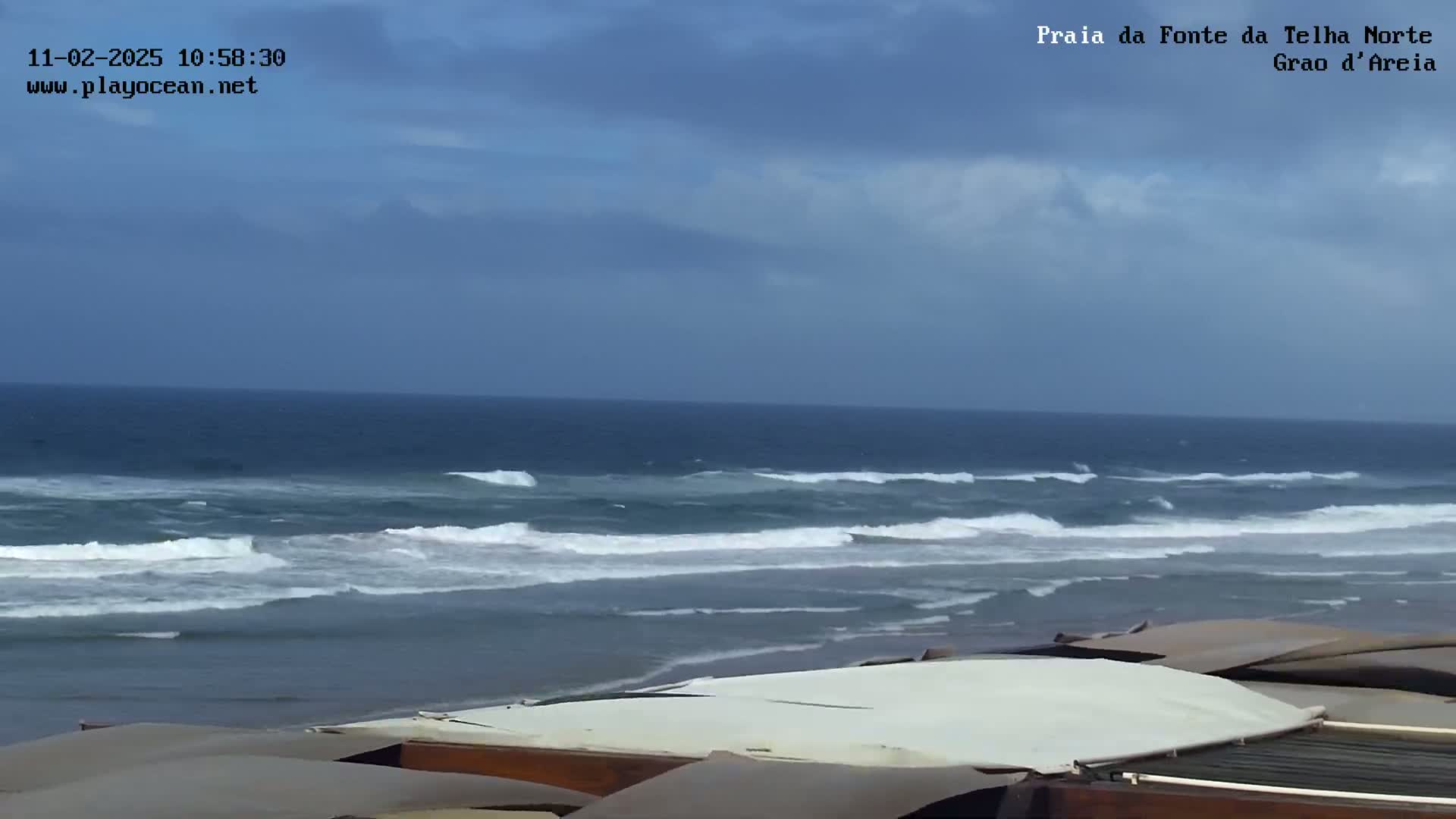 An overcast sky overlooks a beach with moderate waves rolling into shore, partially obscured by the tops of several structures.