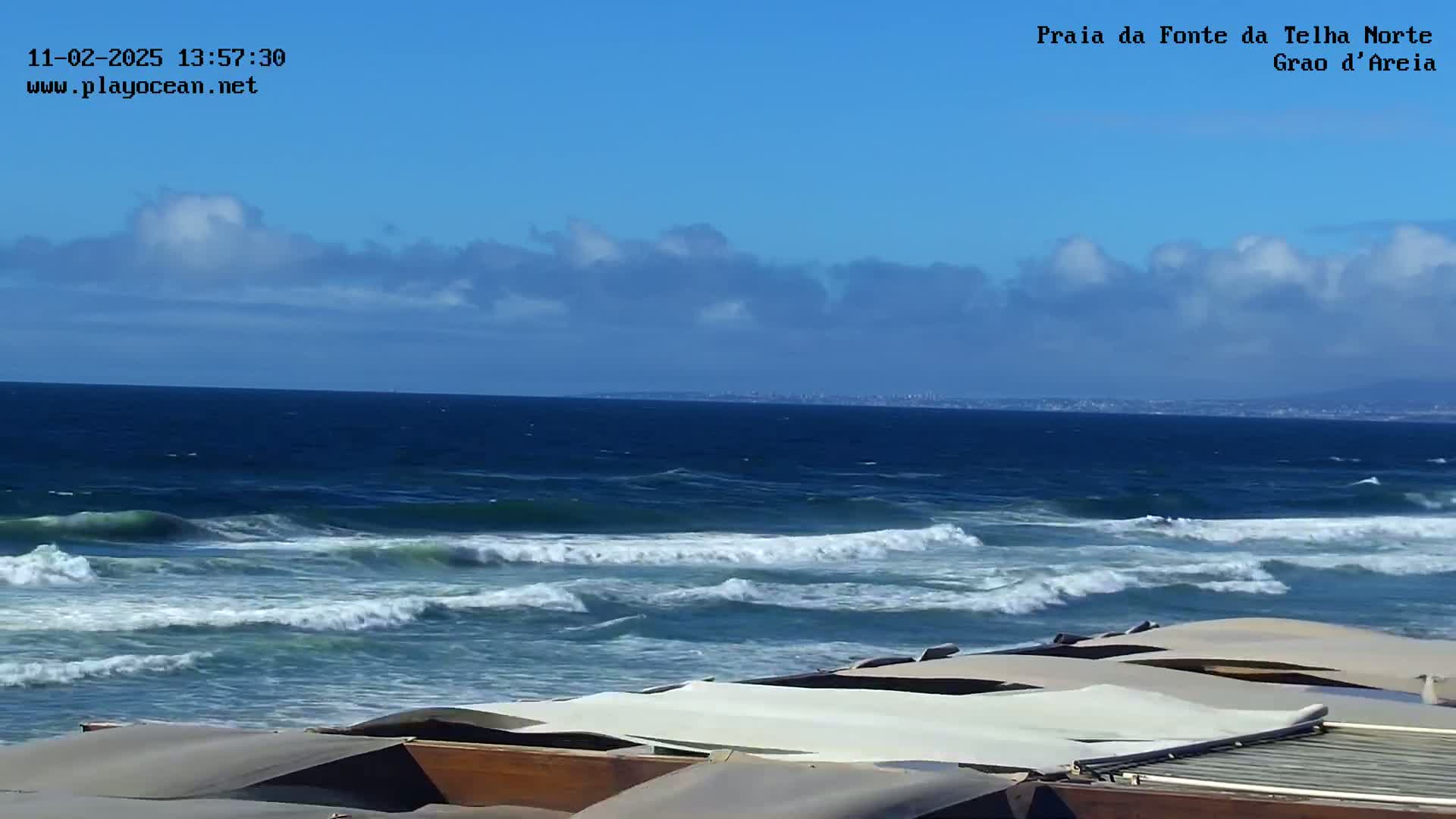 A partly cloudy blue sky overlooks a dark blue ocean with moderate waves crashing onto a shore where several beige canopies are collapsed on wooden structures.