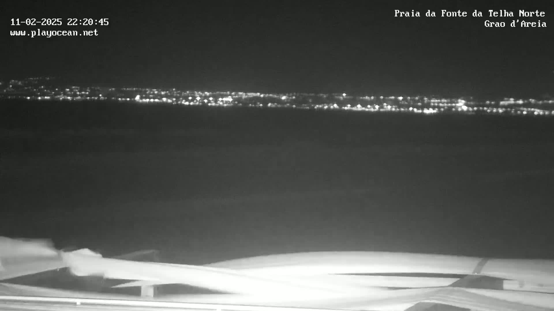 A nighttime, long shot shows a distant city skyline illuminated with lights across a dark body of water.