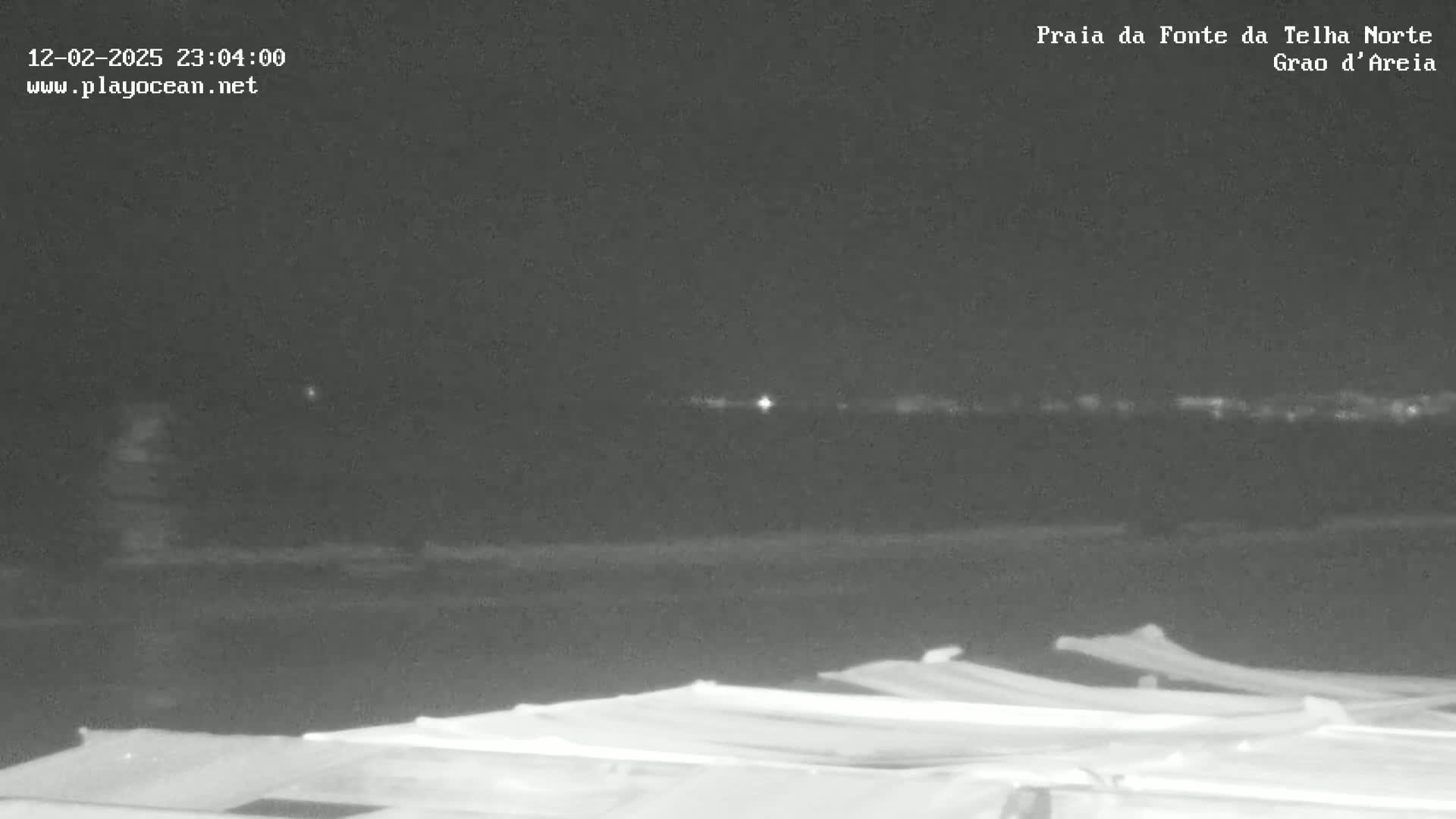 A nighttime, low-light image shows a dark beach with a few indistinct objects in the foreground, possibly beach umbrellas or coverings, and a dark horizon line with a few distant lights visible.