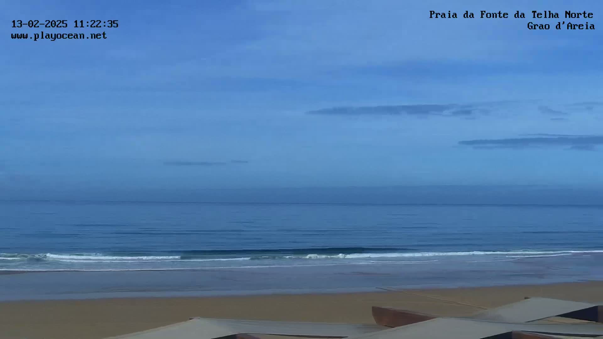 A tranquil ocean scene under a clear, pale blue sky shows calm waves lapping a sandy beach.