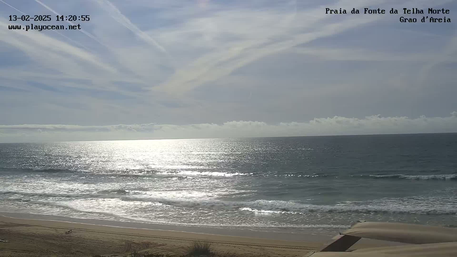A sandy beach with gently rolling waves under a mostly clear sky with a few wispy clouds.