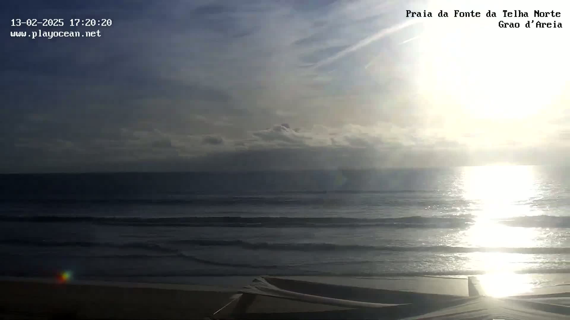 A mostly cloudy, sunny day at a beach shows a calm ocean with small waves lapping the shore.