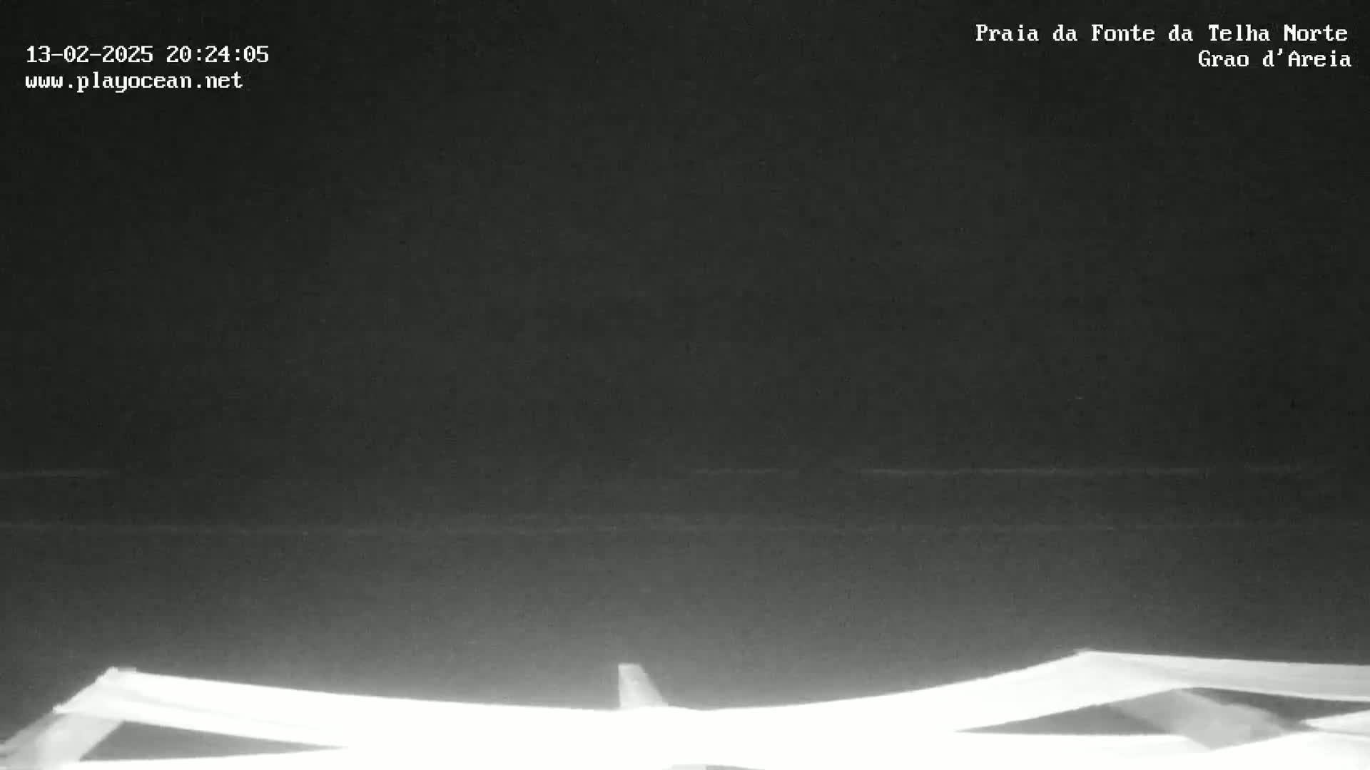 A nighttime, low-light view of a dark beach with calm water and the tops of several beach chairs or loungers visible in the foreground.