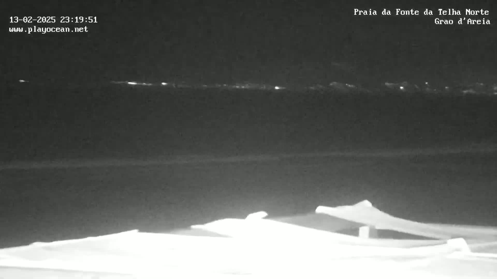 A nighttime, low-light image shows a dark ocean with a faintly visible shoreline in the distance, and in the foreground, several indistinct objects appear to be on the ground.