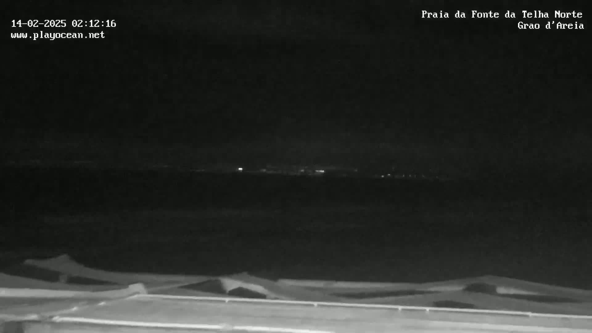 A nighttime, low-light view shows a dark body of water with a distant shoreline faintly illuminated by lights, seen from behind a low structure with a slightly wavy, textured top.