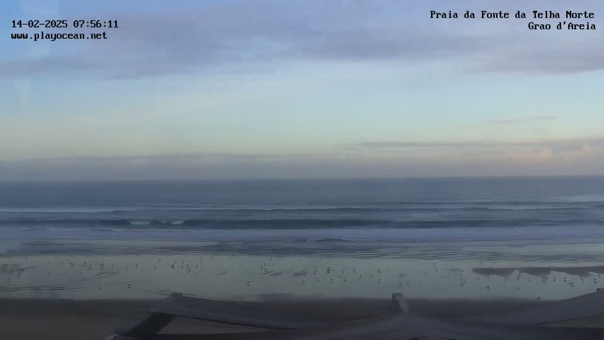 A wide shot of a beach with gently rolling waves under a mostly cloudy, pale sky.