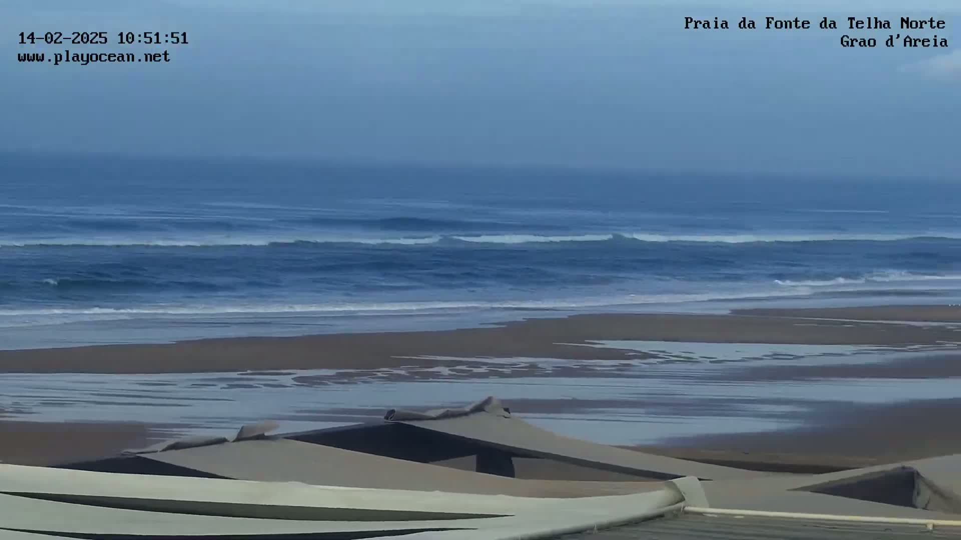A wide sandy beach with gentle waves under an overcast sky is partially obscured by a low structure in the foreground.
