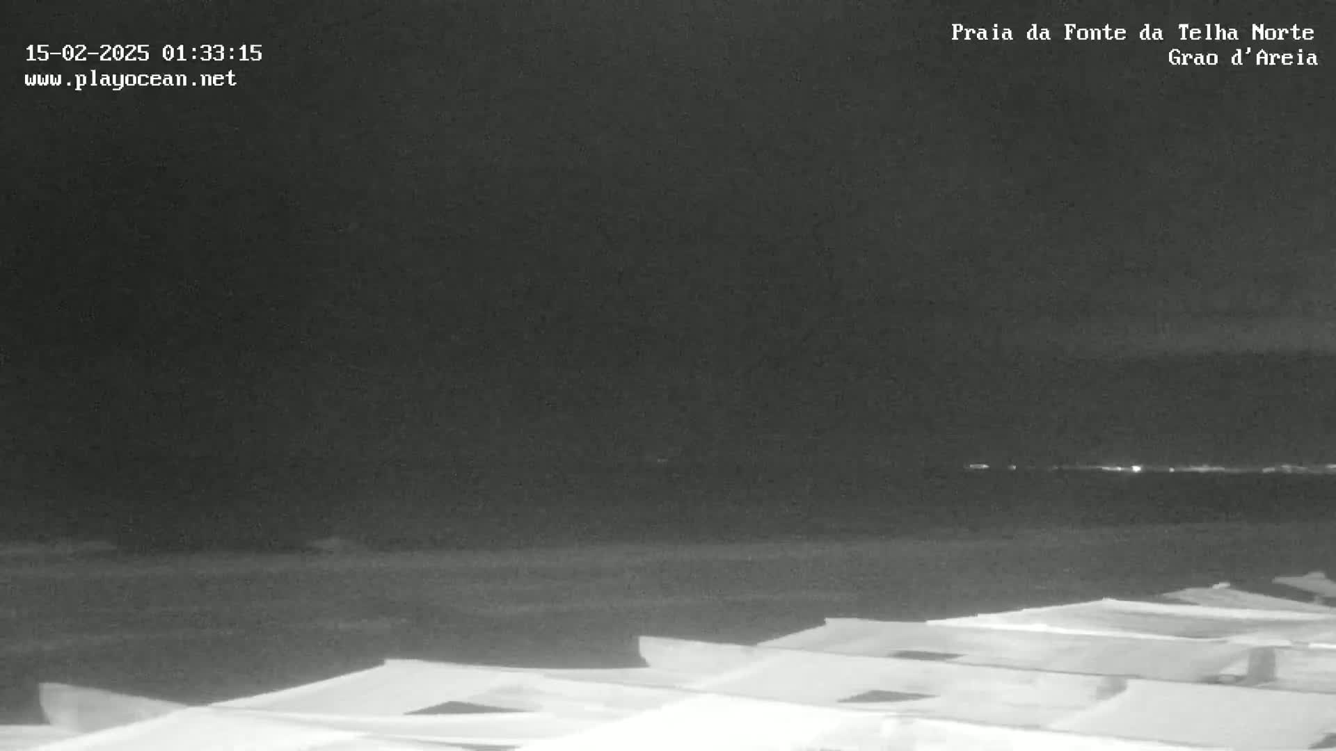 A nighttime, low-light view of a beach shows several boats or similar objects in the foreground, with a dark body of water and a faint, distant light source visible on the horizon.