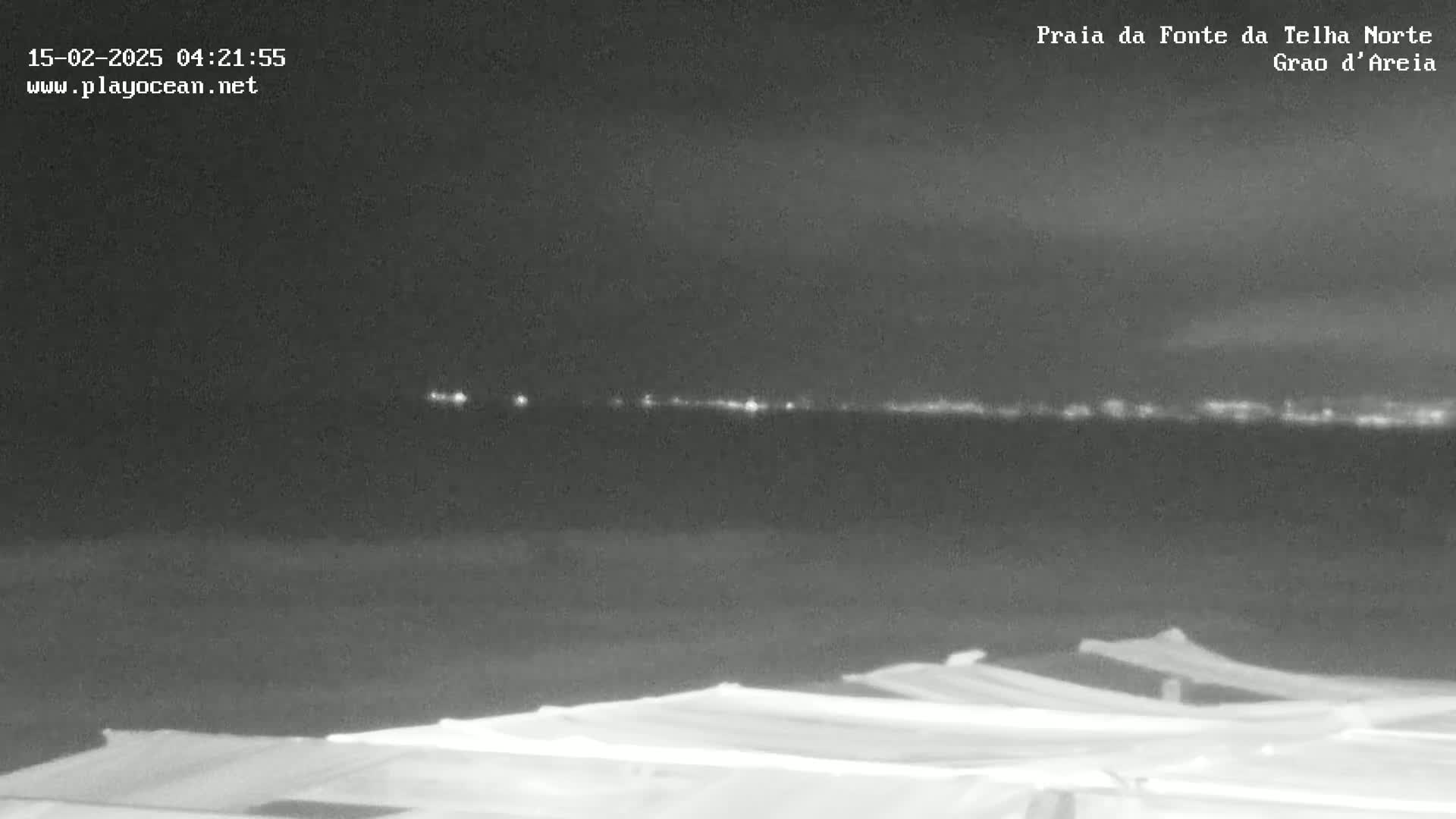 A nighttime view shows a dark beach with several covered structures in the foreground and a distant, faintly lit shoreline under a cloudy sky.