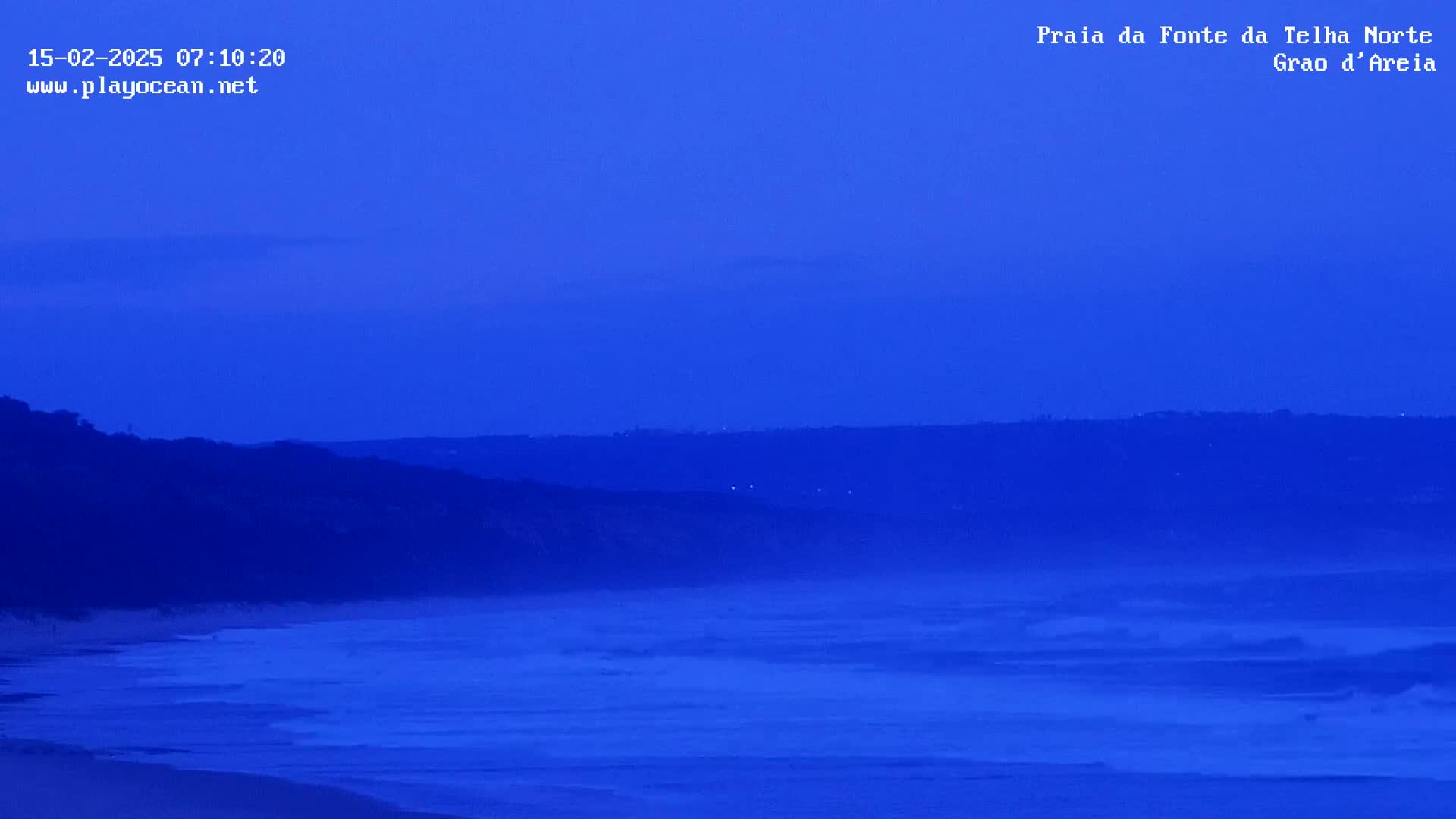 Under a twilight blue sky, a dark coastline meets a dark blue ocean with small waves.