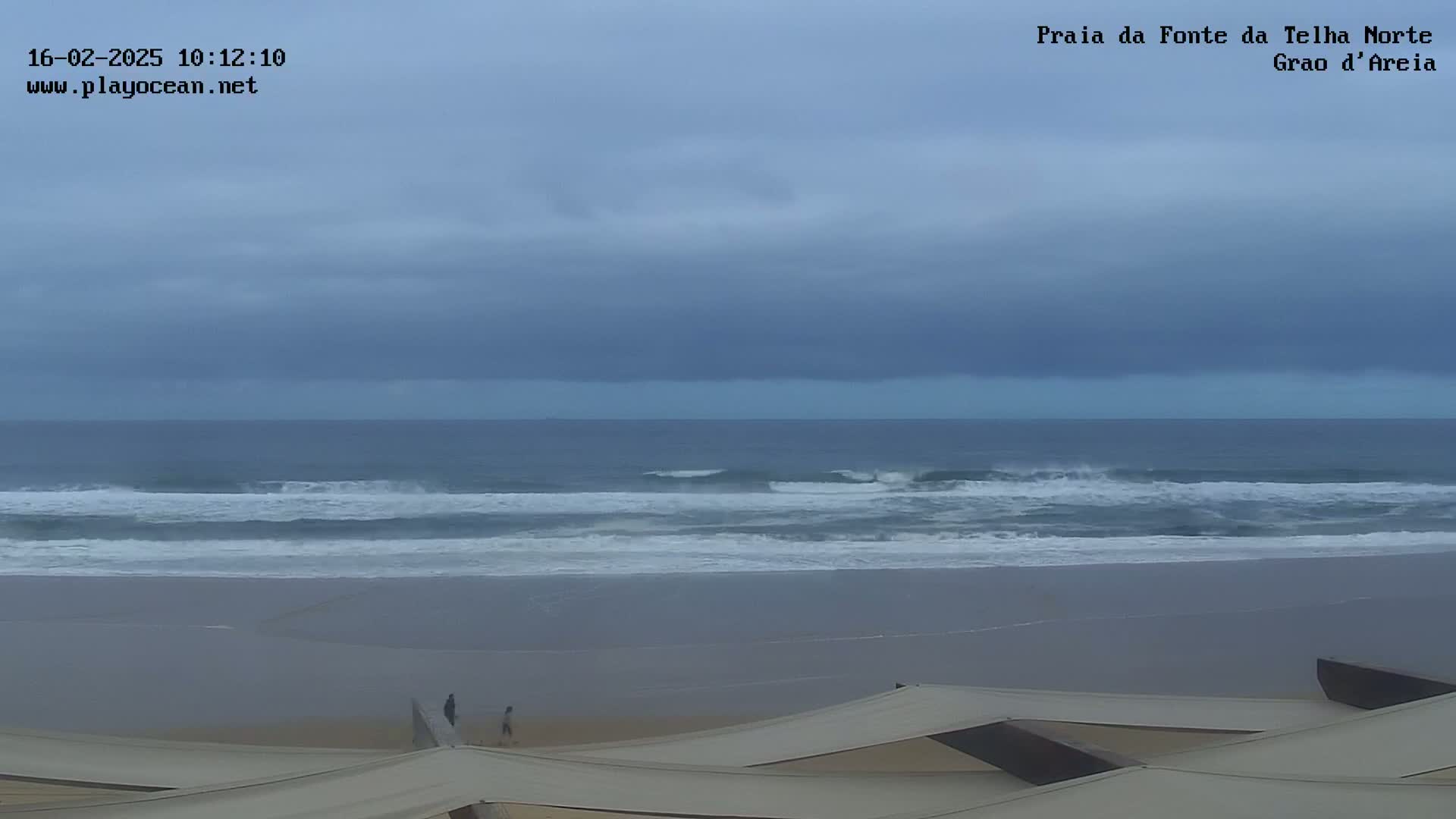 A wide shot of an overcast beach scene shows a grey sky, moderate waves, and a few people walking on a mostly empty sandy beach.