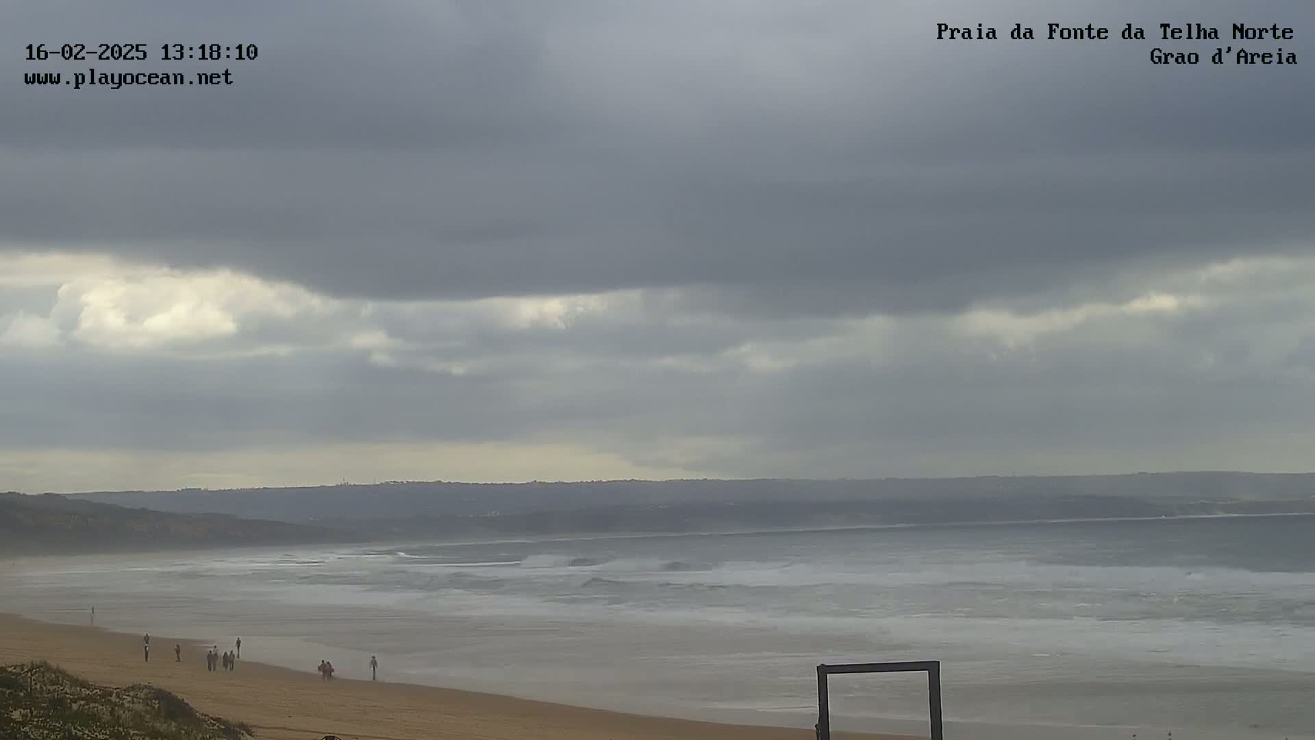 A sandy beach with a few people walking along the shore under an overcast sky, with gentle waves rolling into the ocean.