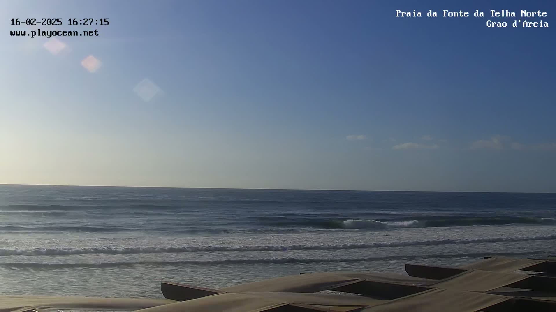 A mostly clear blue sky overlooks a calm ocean with small waves lapping a sandy shore where several brown structures are partially visible in the foreground.