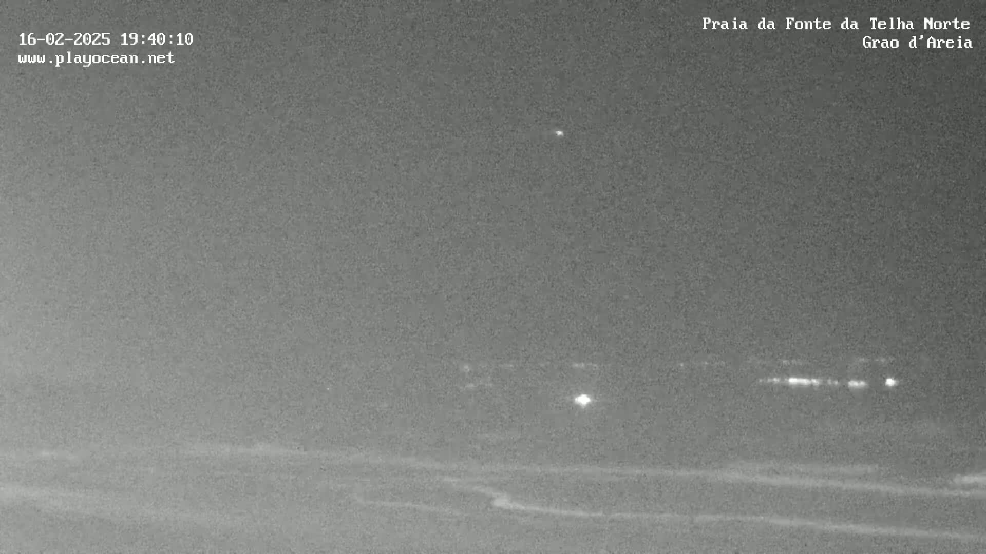 A nighttime, low-light view of a beach with a distant line of lights, possibly a town or village.
