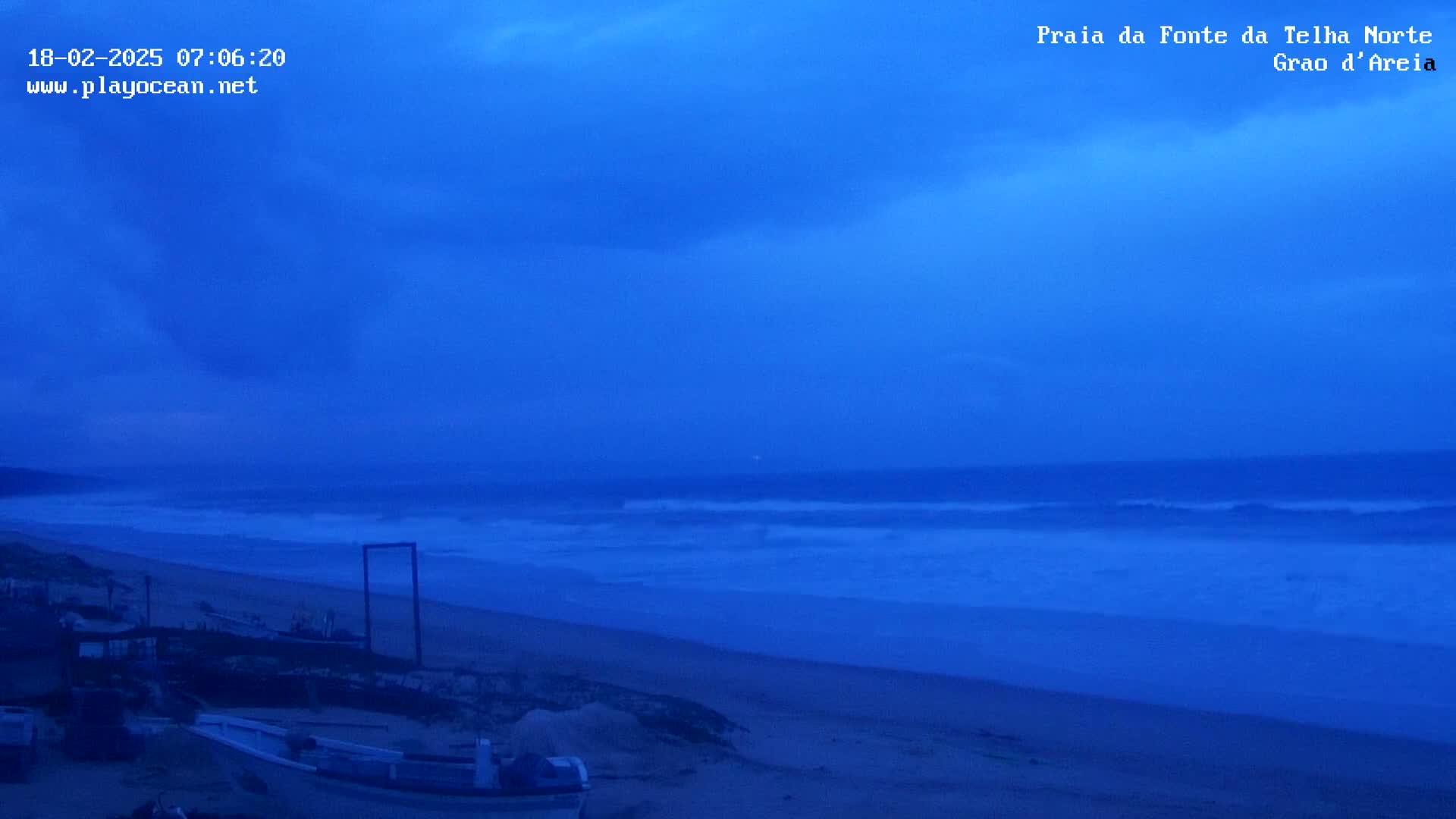 A sandy beach with several small boats pulled ashore is visible under a twilight sky, with dark clouds and a somewhat turbulent ocean in the background.