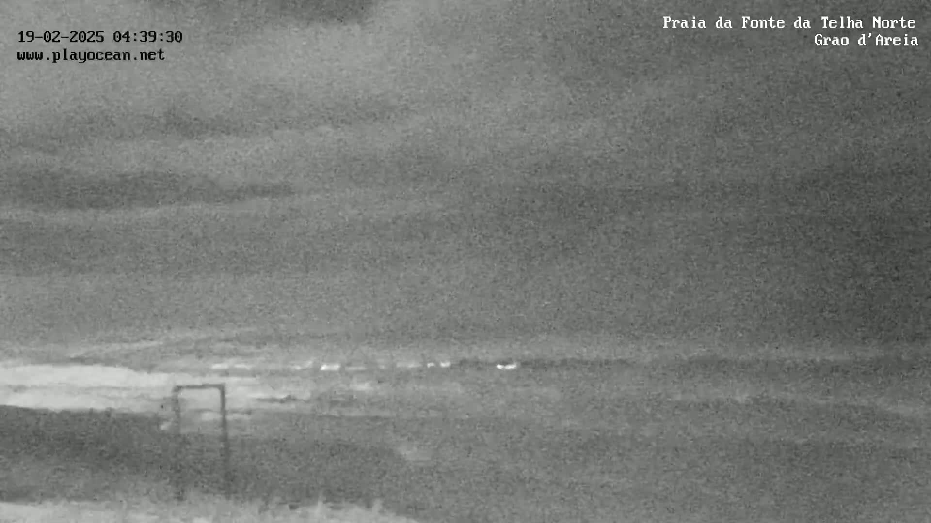 A nighttime, grayscale image shows a dark beach and ocean under an overcast sky.