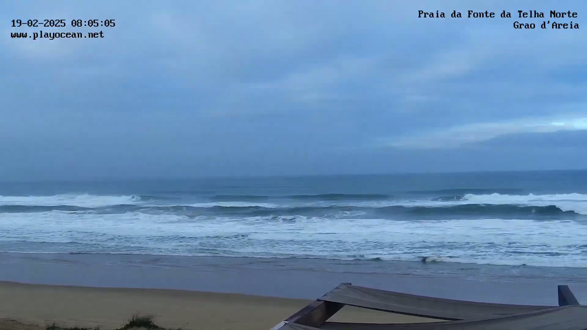 A beach with moderate waves under an overcast sky is seen from a slightly elevated vantage point.