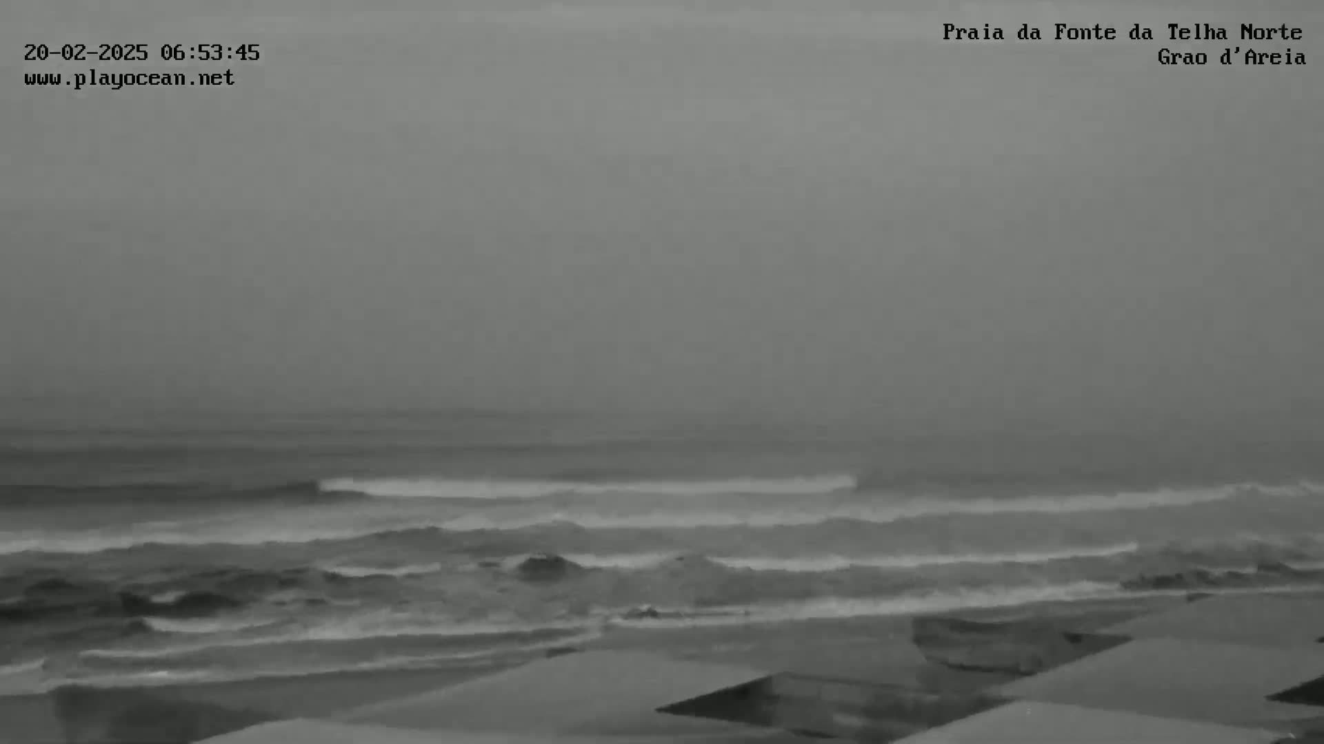 A grayscale image shows an overcast day at a beach with moderate waves breaking near the shore.