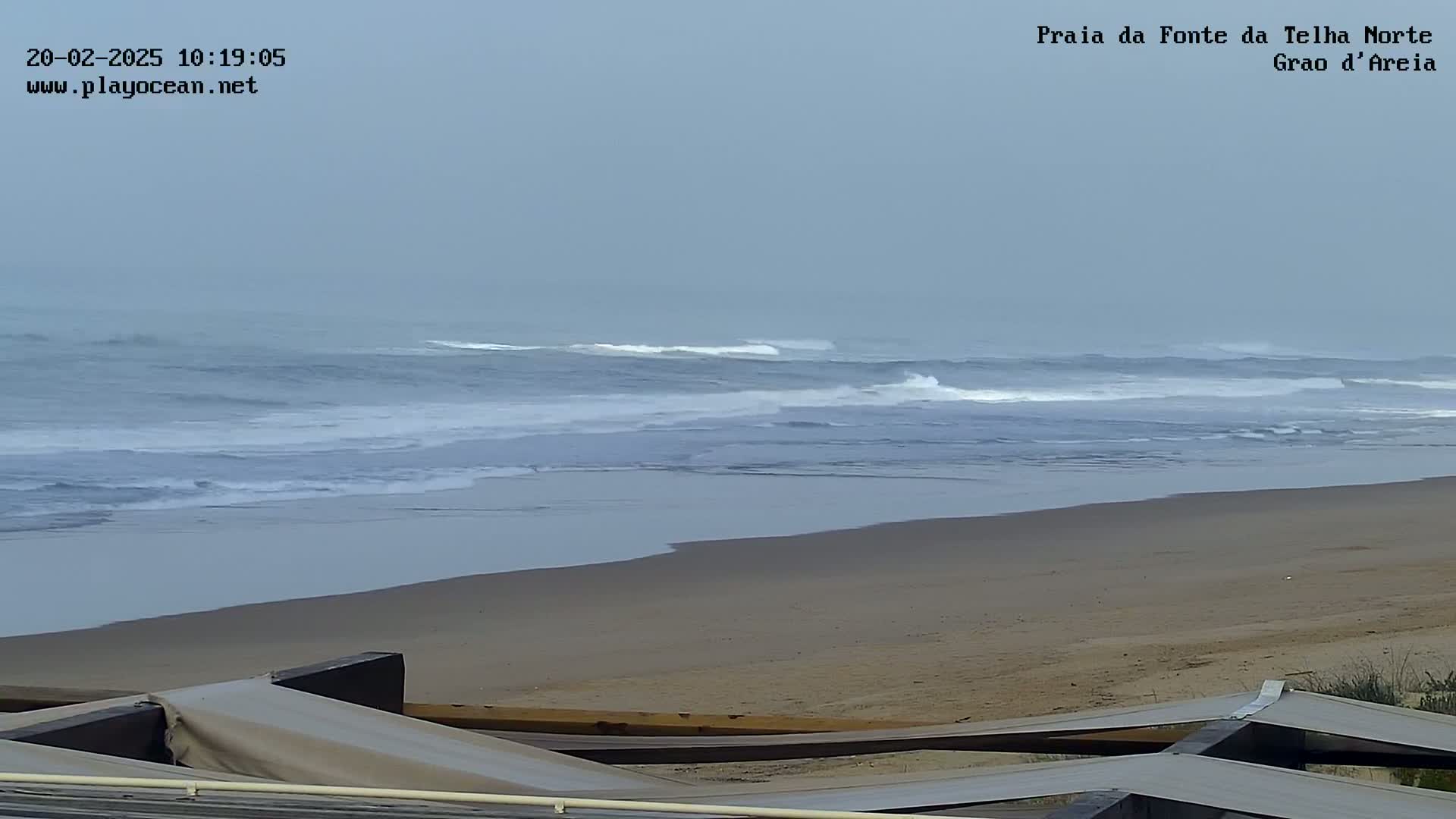 A sandy beach with gentle waves under an overcast sky is partially obscured by a structure in the foreground.