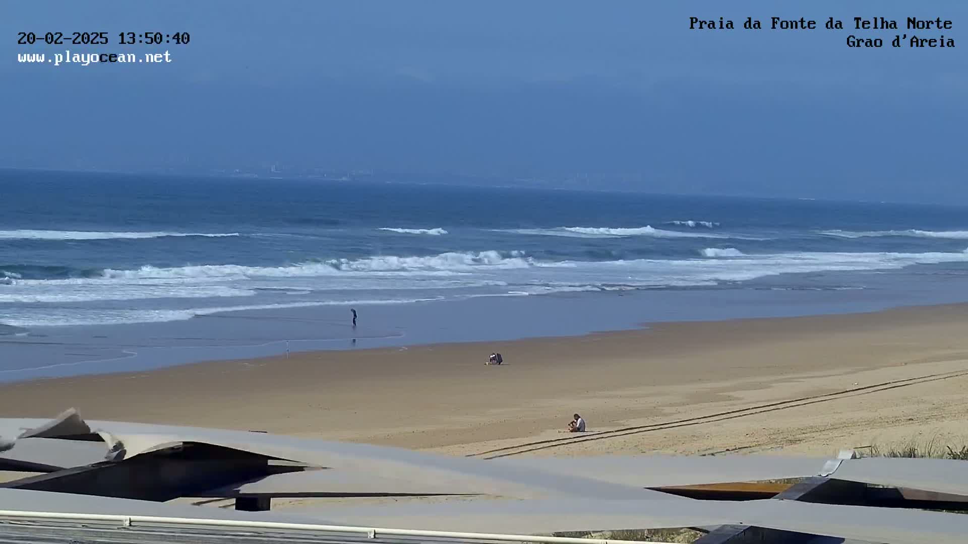 A sandy beach with small waves rolling onto the shore on a clear, sunny day, with a few people visible on the beach and a distant view of land on the horizon.