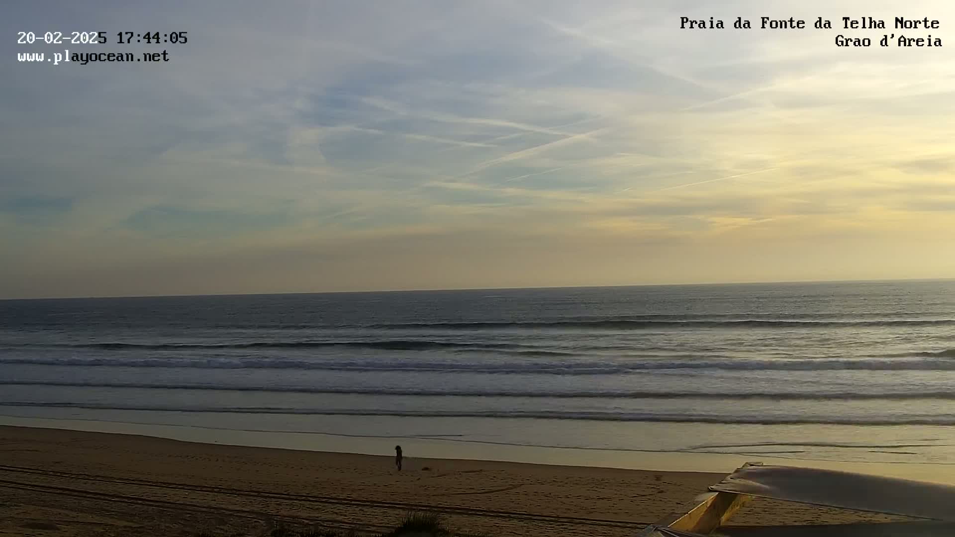 Under a partly cloudy sunset sky, gentle waves roll onto a sandy beach where a lone person walks near the water's edge.