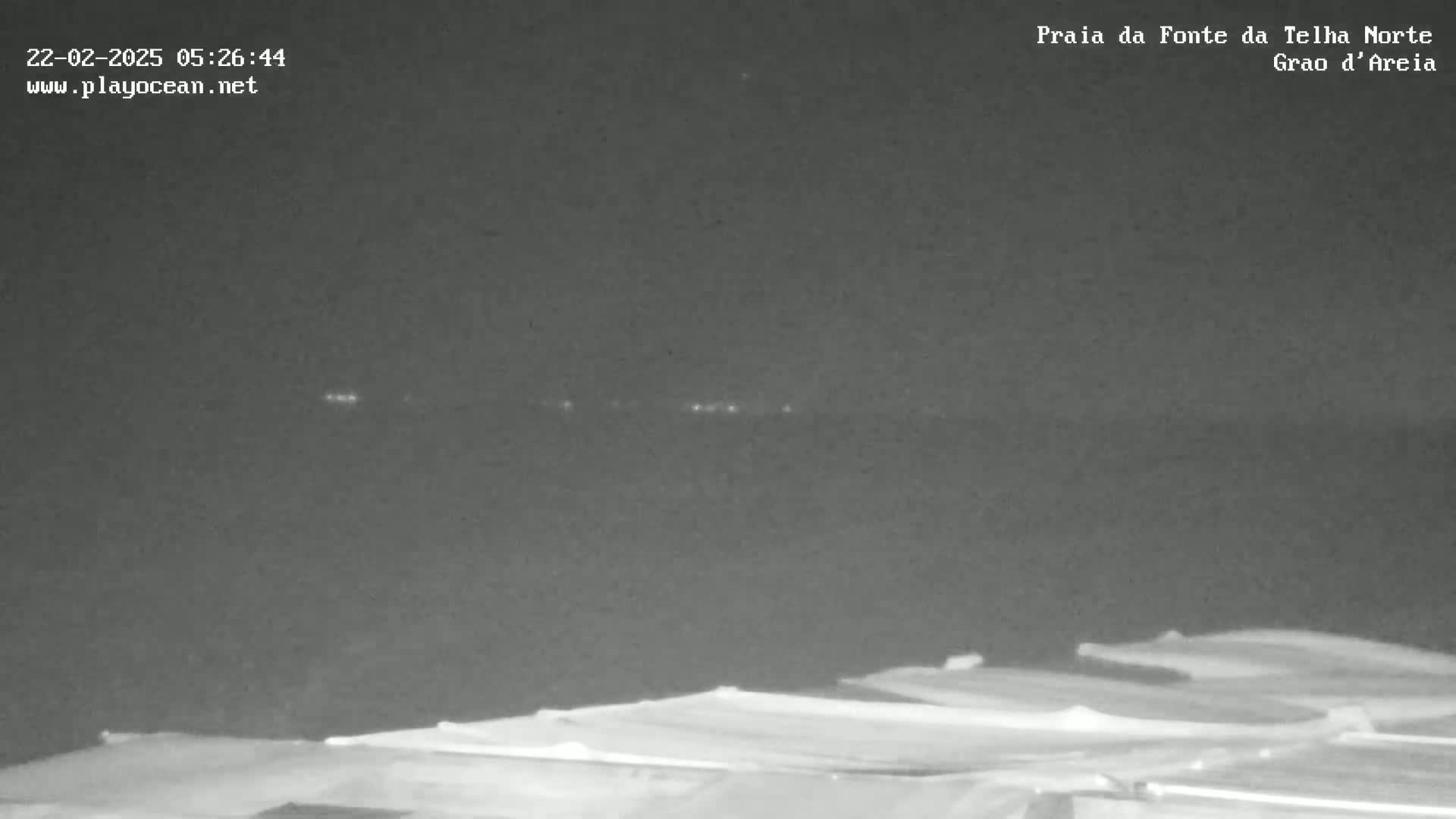 A nighttime, low-light image shows a dark body of water with faint lights on the horizon, and in the foreground, a series of low-lying, flat structures.