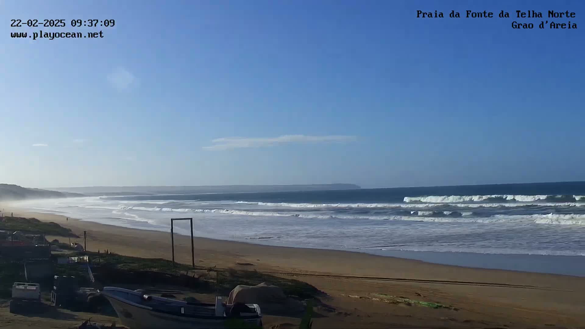 A sandy beach with moderate waves under a clear blue sky stretches towards a distant coastline.
