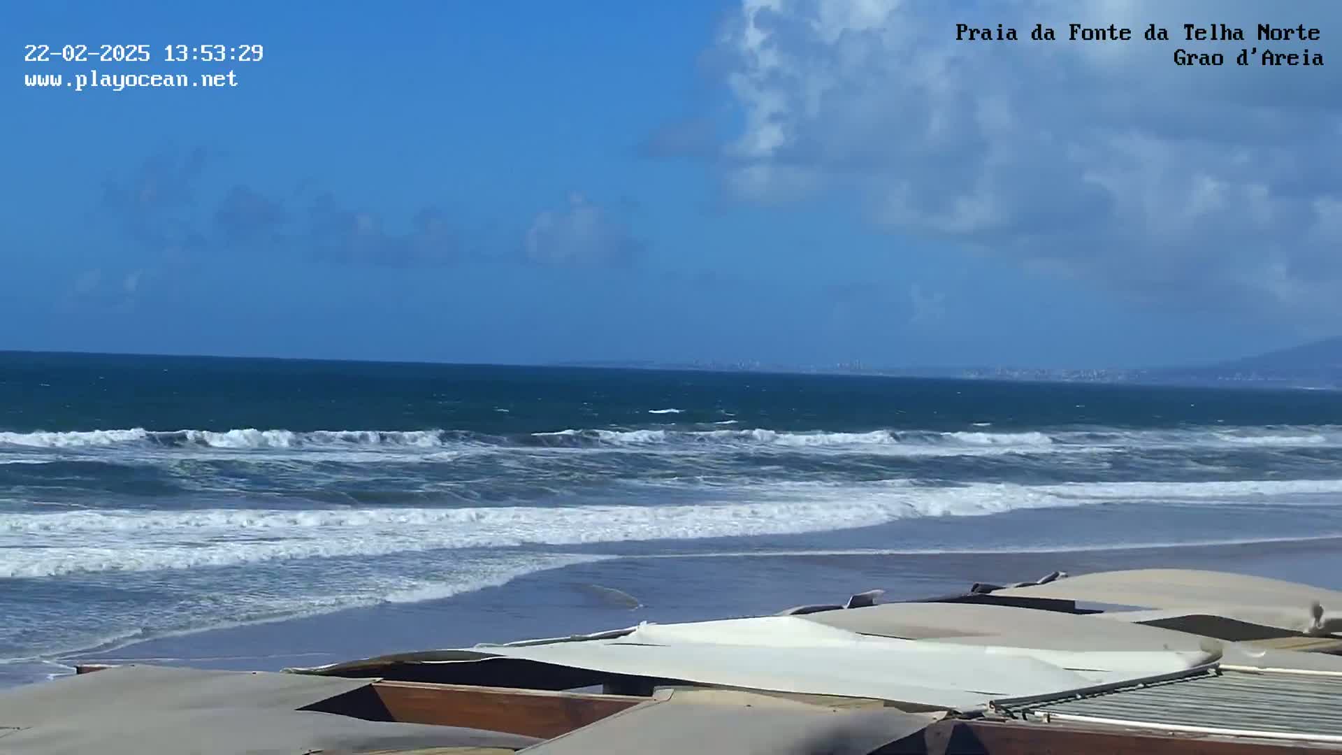 A partly cloudy blue sky overlooks a beach with moderate waves lapping the shore, where several folded awnings are situated in the foreground.