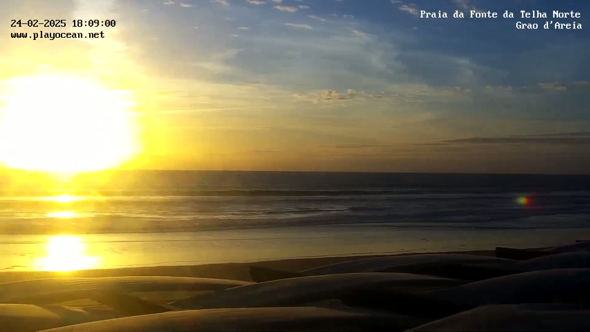 A bright sunset over a calm ocean beach with several dark, covered objects on the sand.
