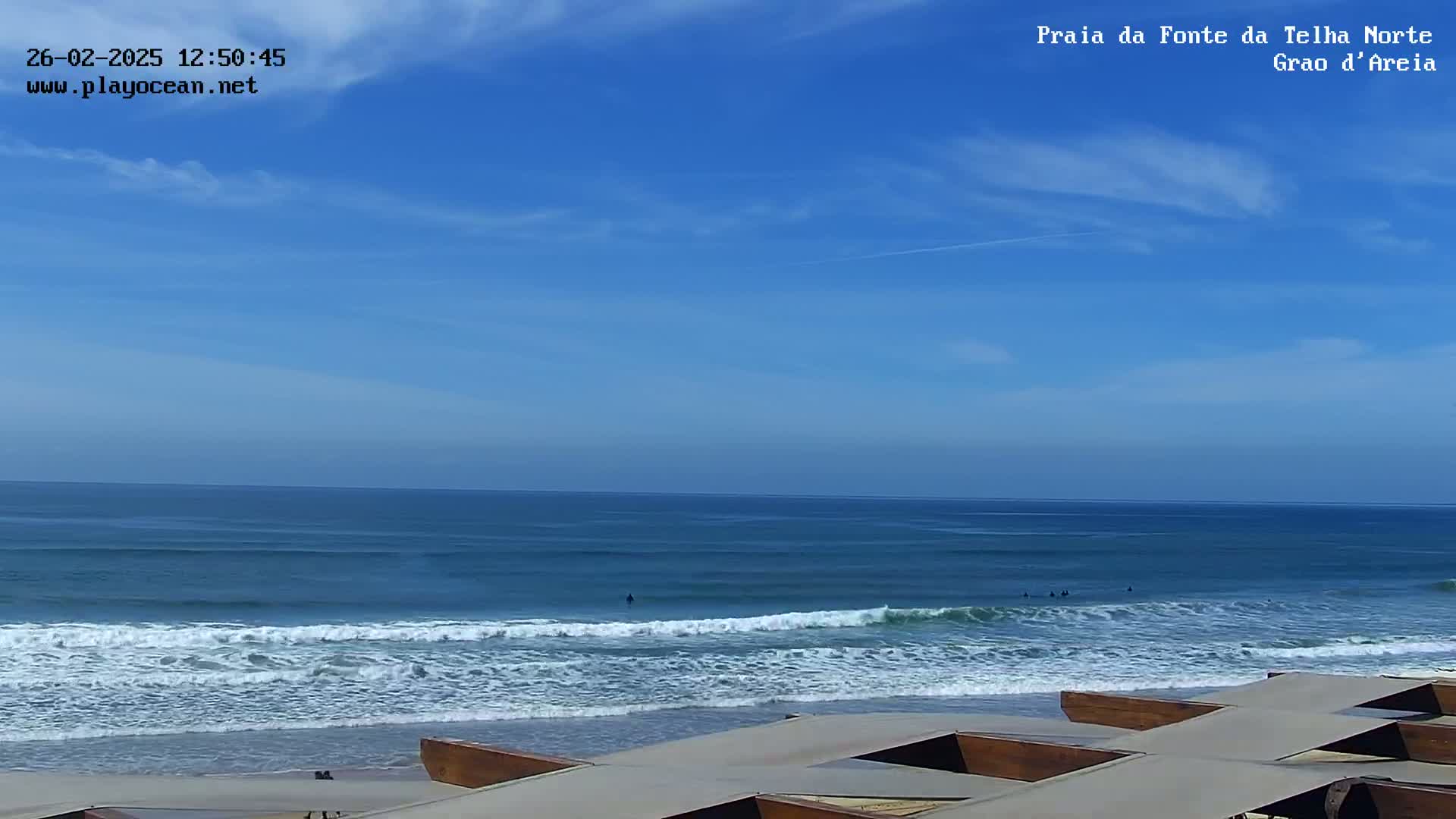 Under a mostly clear blue sky, a tranquil ocean scene shows several surfers in the waves that gently roll onto a sandy beach with wooden structures in the foreground.