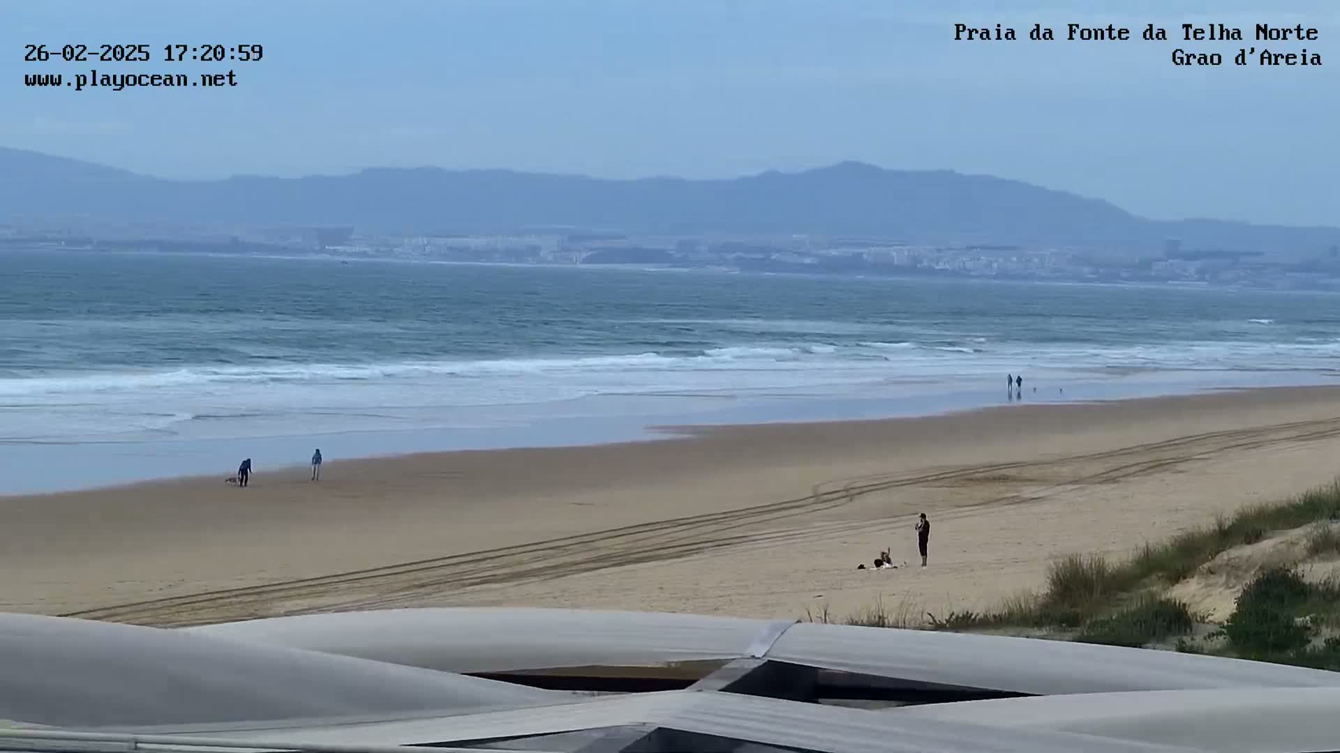 A wide sandy beach with a few people on it stretches toward a calm ocean under a clear sky, with distant mountains visible on the horizon.
