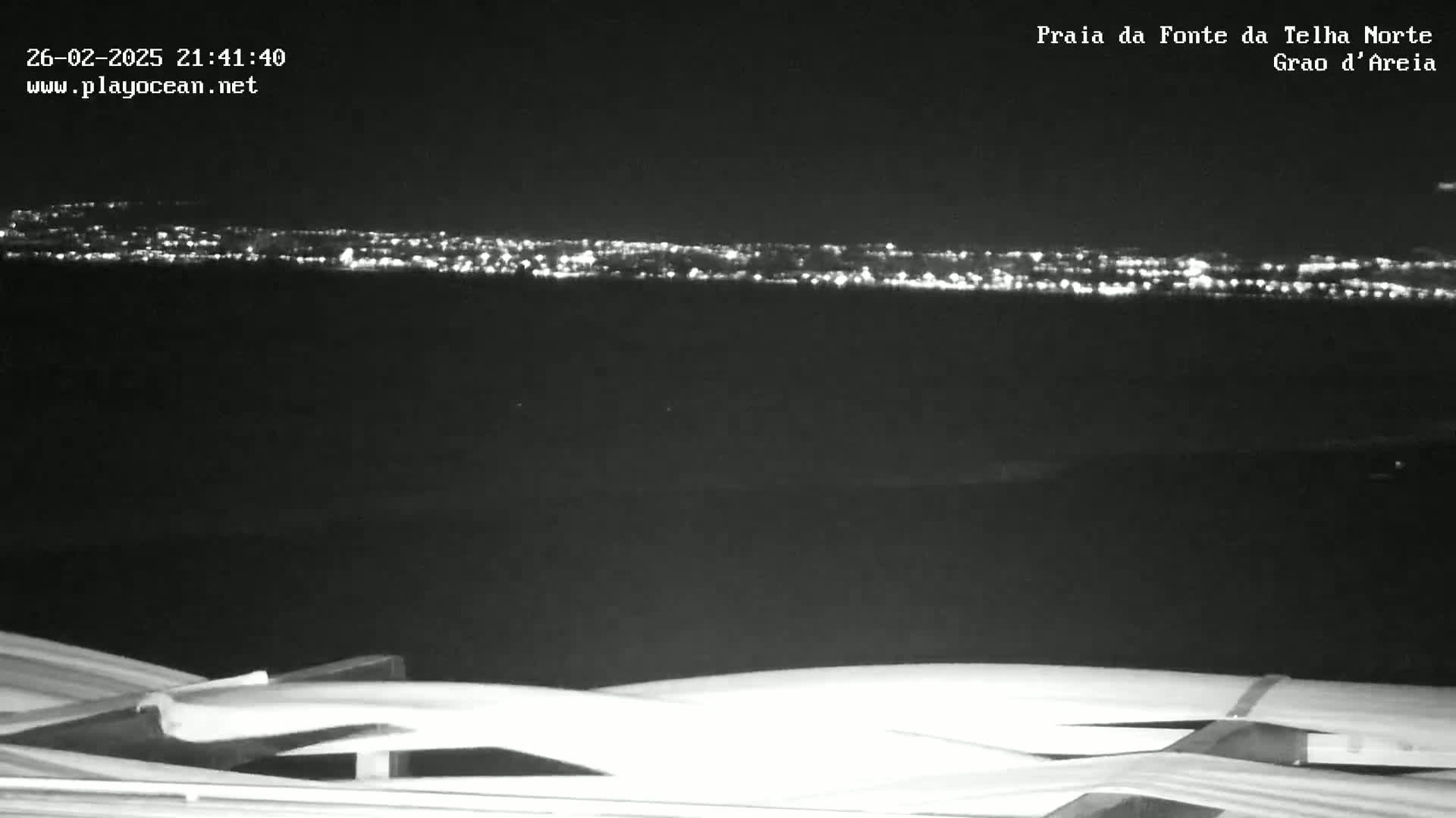 A nighttime, long-range view shows a distant city skyline illuminated by lights, with a dark foreground featuring a curved, white structure in the bottom of the frame.