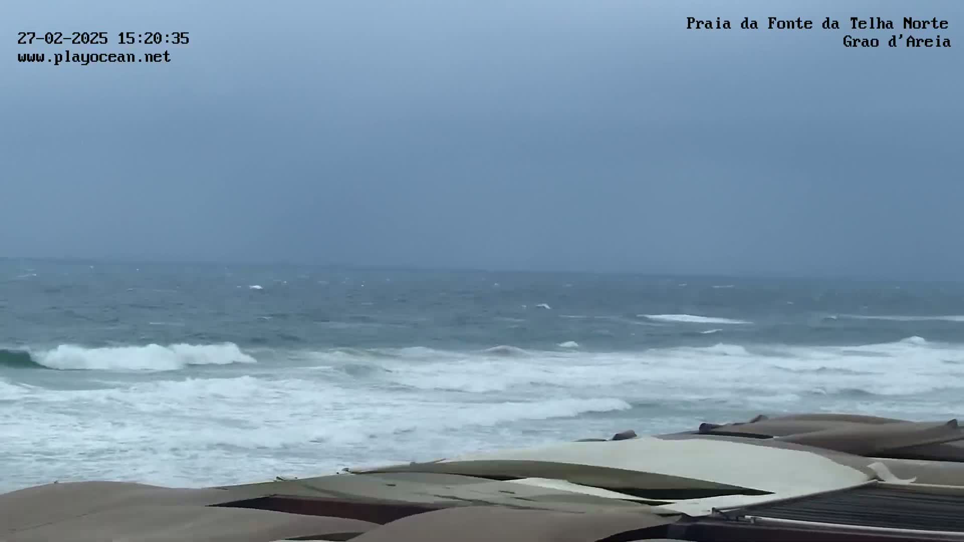 A partly cloudy day shows choppy ocean waves crashing on a beach where several closed-up beach umbrellas or sunshades sit in the foreground.