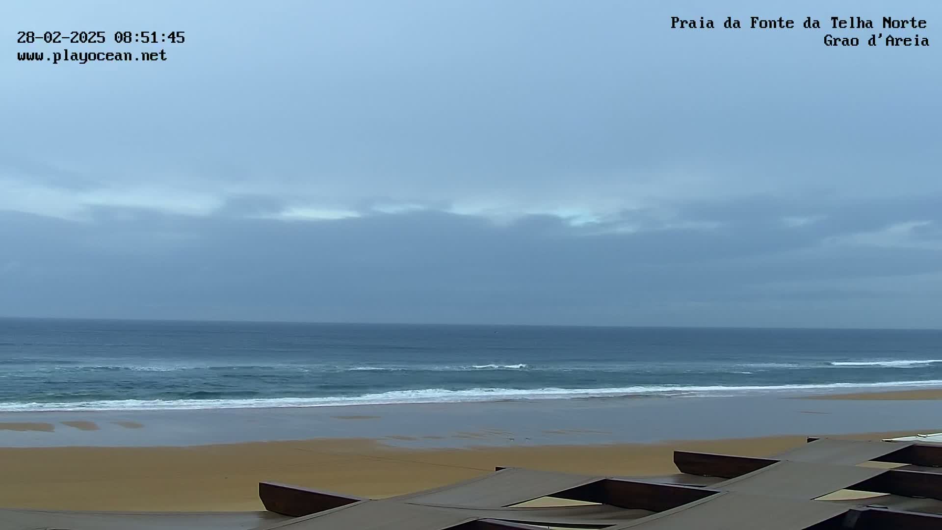 A wide sandy beach with gentle waves under a cloudy sky is viewed from a slightly elevated position, showing a partial view of some beach furniture in the foreground.