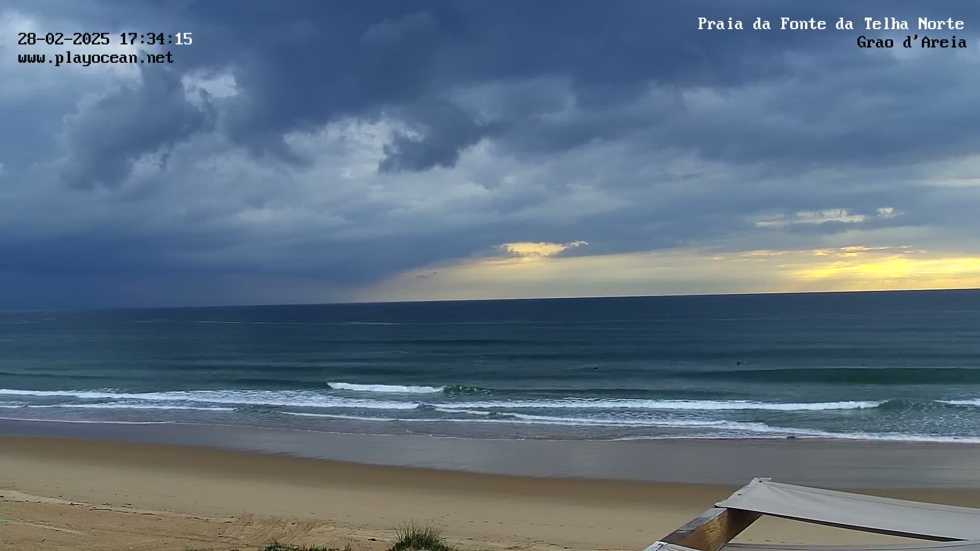 A sandy beach with gentle waves rolls into a dark, overcast ocean under a mostly cloudy sky with a hint of yellow light on the horizon.
