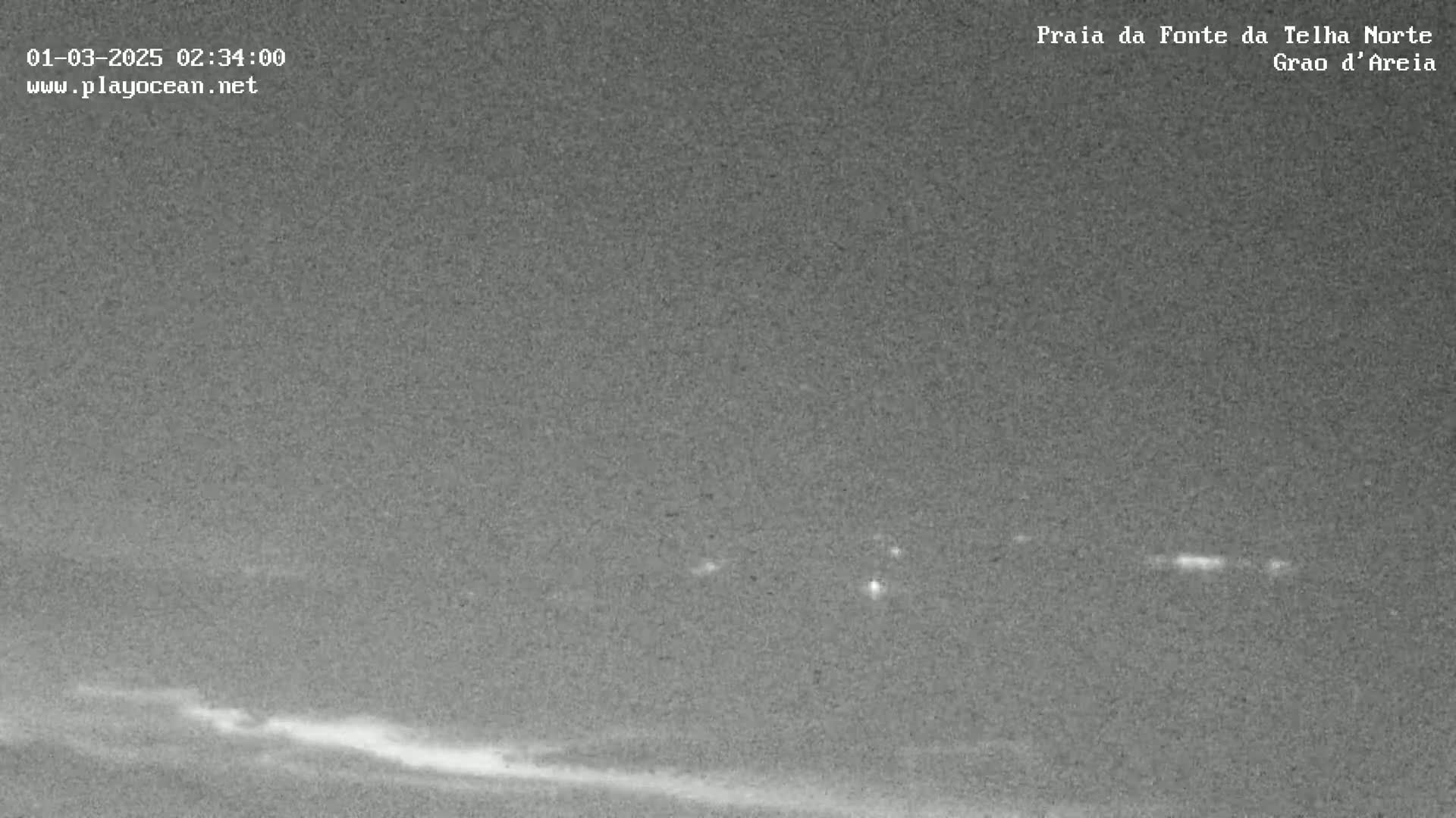 A nighttime, low-light view shows a dark beach and ocean with a faint line of light on the horizon.