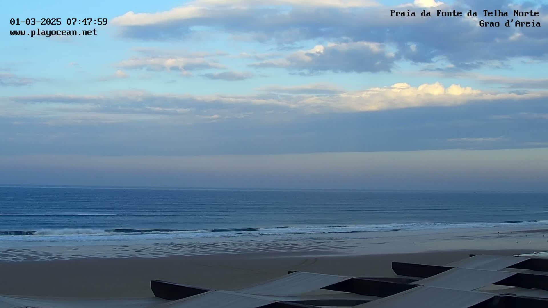 Under a mostly cloudy sky, a wide sandy beach slopes down to a calm ocean with small waves rolling onto the shore.