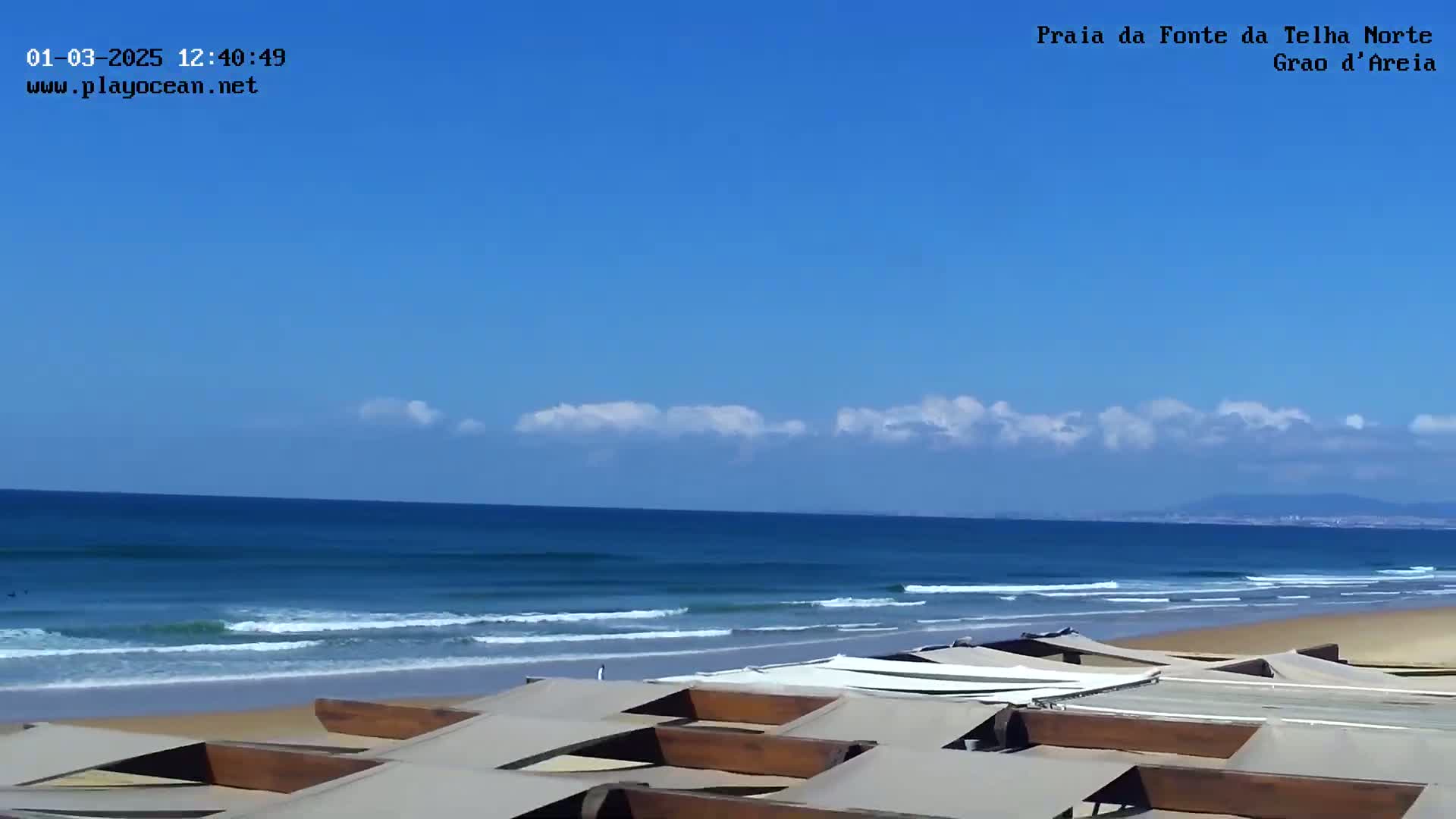 A sandy beach with small waves under a mostly clear blue sky is viewed from behind a row of beach umbrellas.